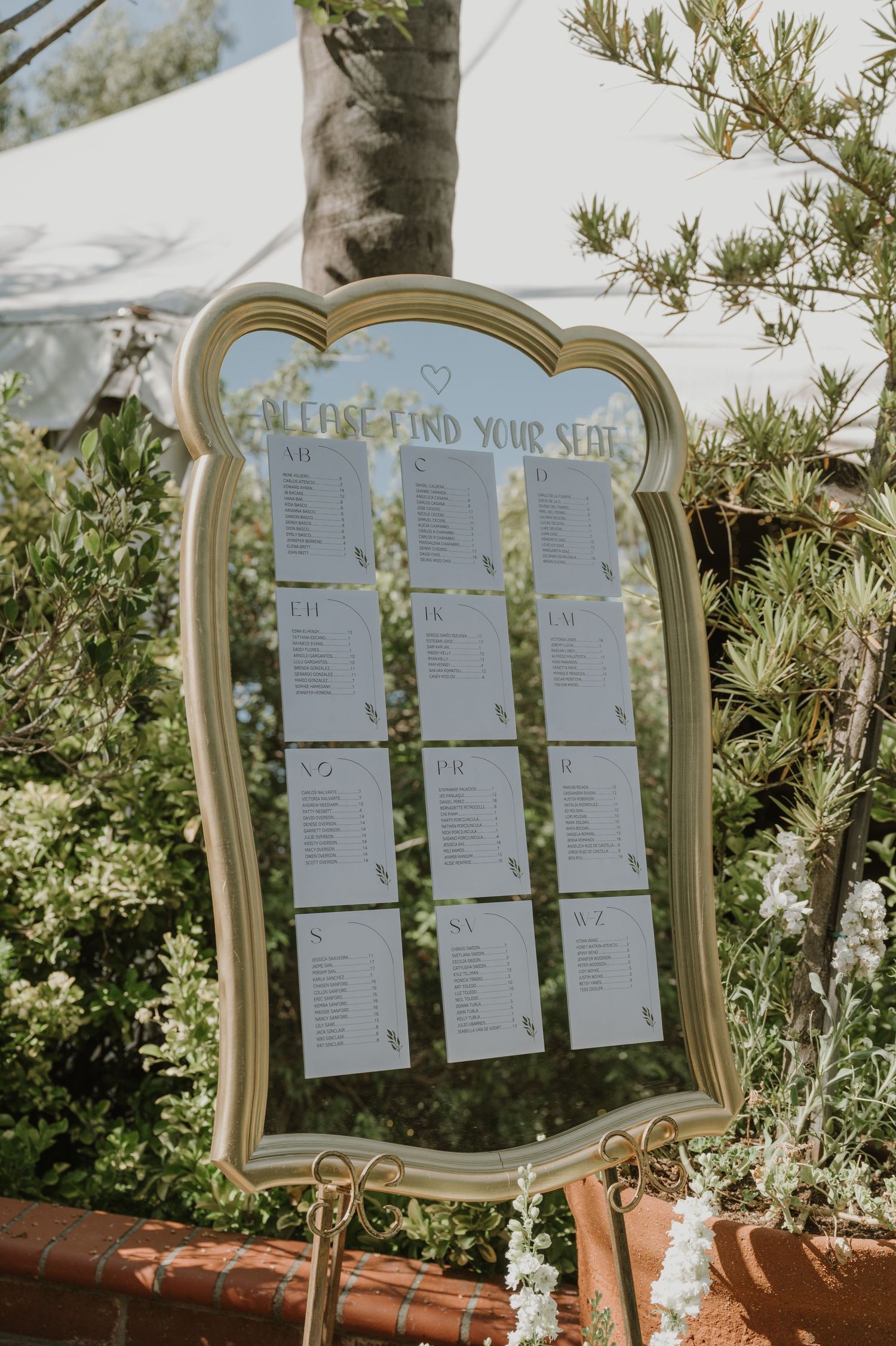 Elegant gold mirror seating chart display at outdoor wedding reception surrounded by lush greenery.