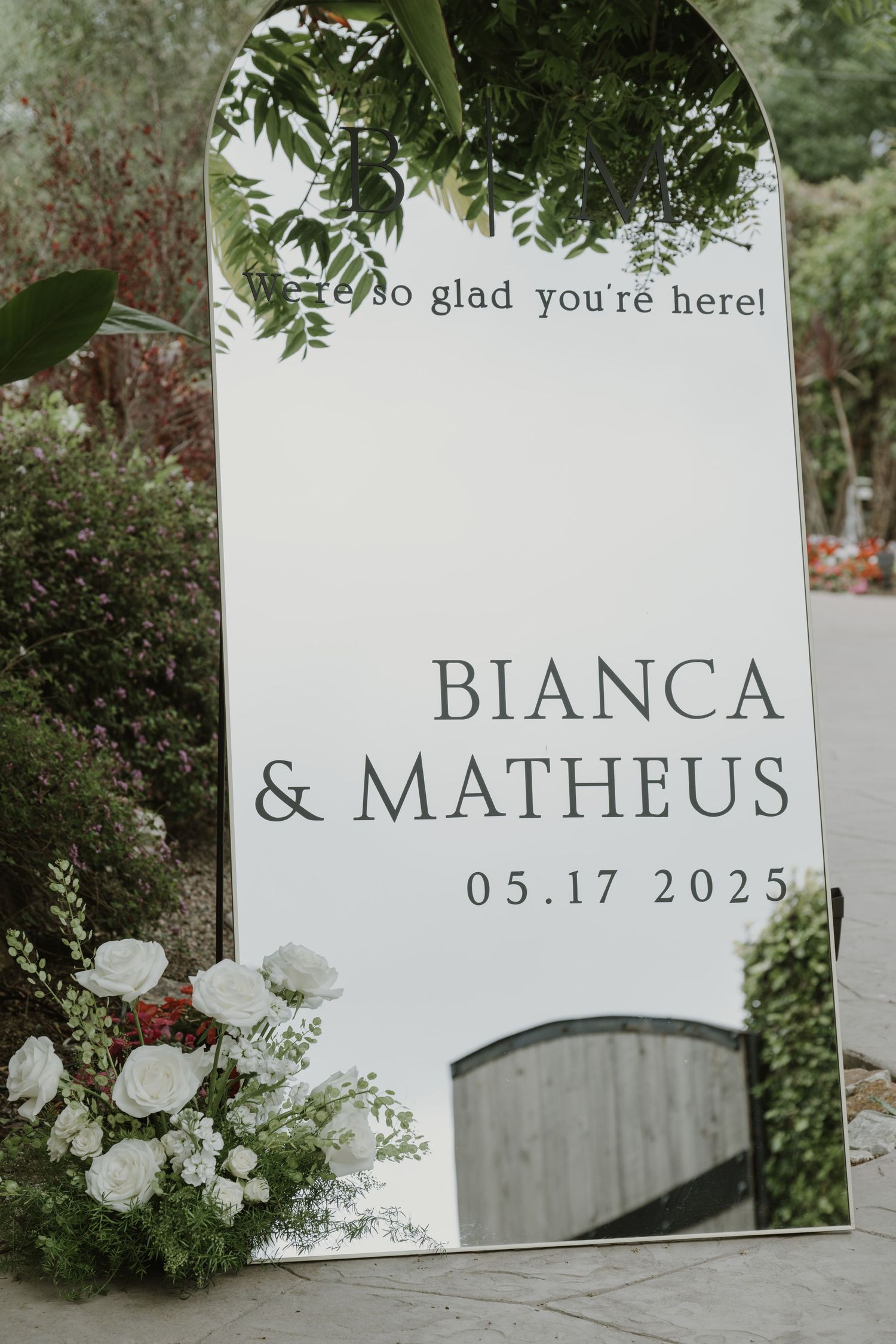 Elegant wedding welcome mirror sign for Bianca and Matheus dated 05.17.2025, adorned with white floral arrangements outdoors.