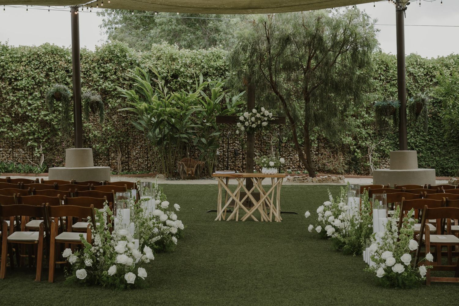 Elegant outdoor wedding ceremony setup with white floral arrangements, wooden chairs, and lush green hedges backdrop.