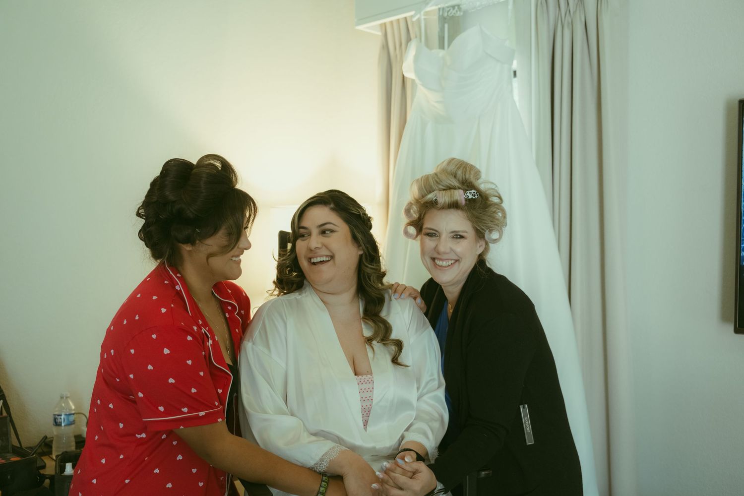 Bride laughing with two bridesmaids in robes during wedding preparations, with bridal gown hanging in background.
