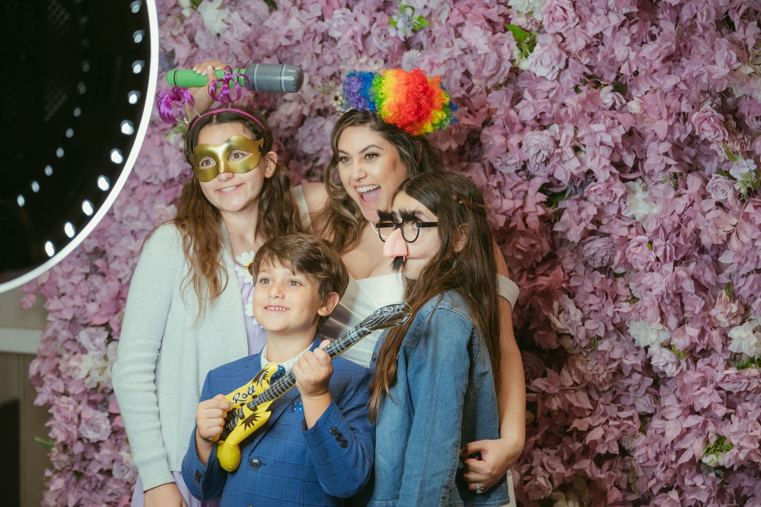 Family of four posing with fun props in front of a floral photo booth wall with pink flowers.