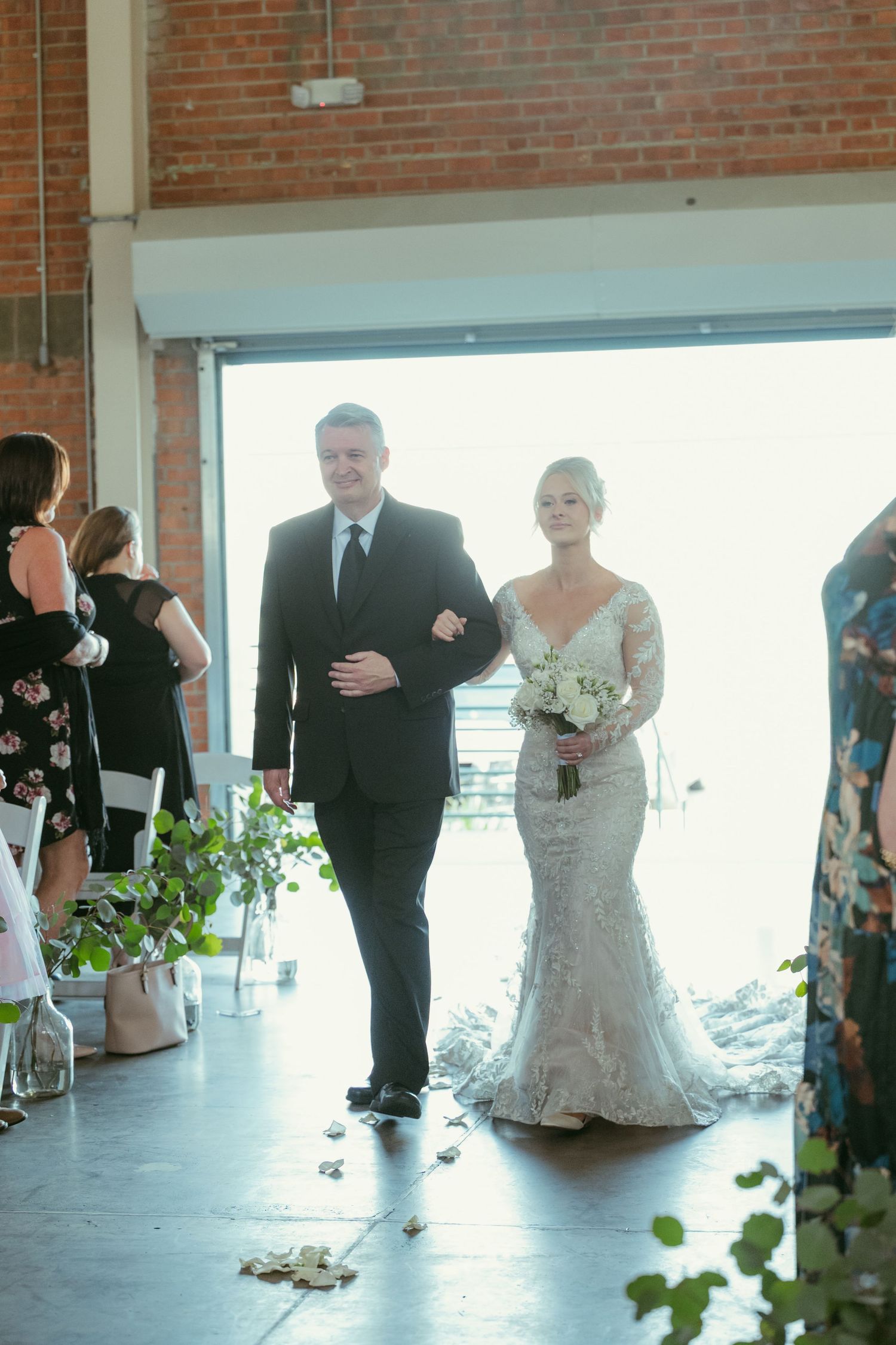 Bride in lace gown walks down aisle with escort at industrial brick wedding venue.