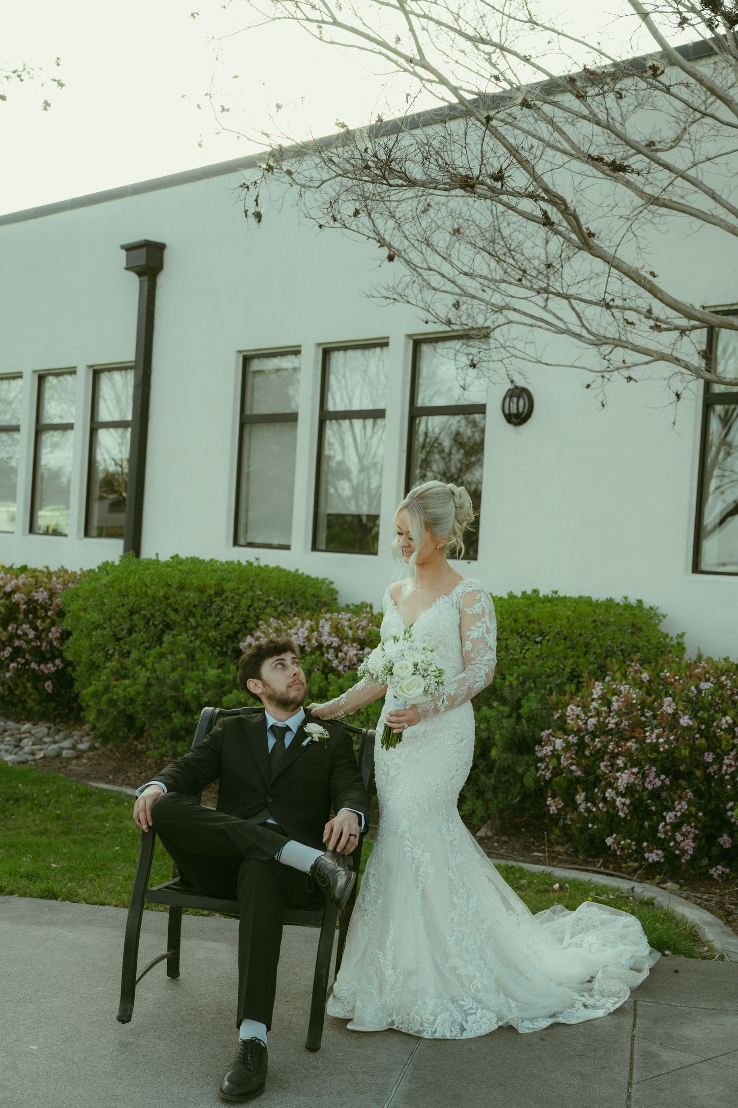 Groom sits on chair gazing up at blonde bride in lace mermaid gown outside white venue.