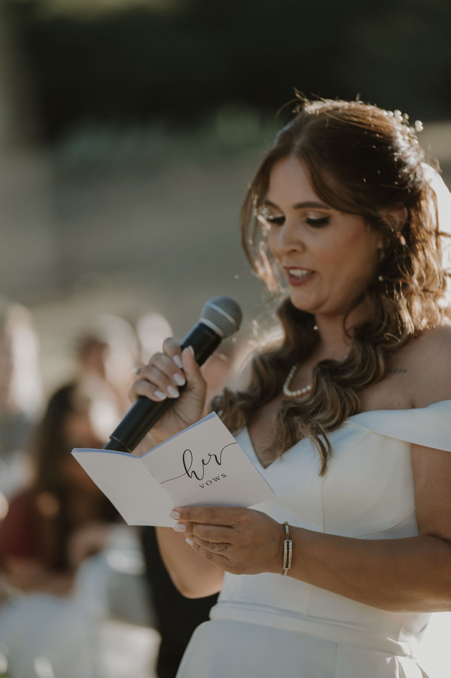 Bride reading her vows from a white booklet while holding a microphone at an outdoor wedding ceremony.