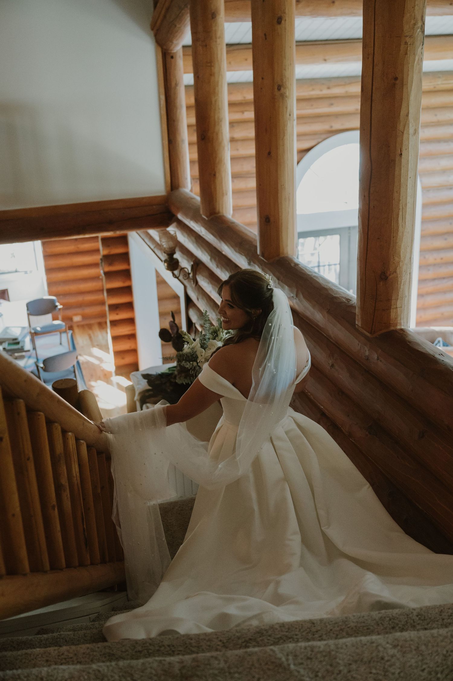 Bride in elegant backless wedding gown with veil descending rustic log cabin staircase.