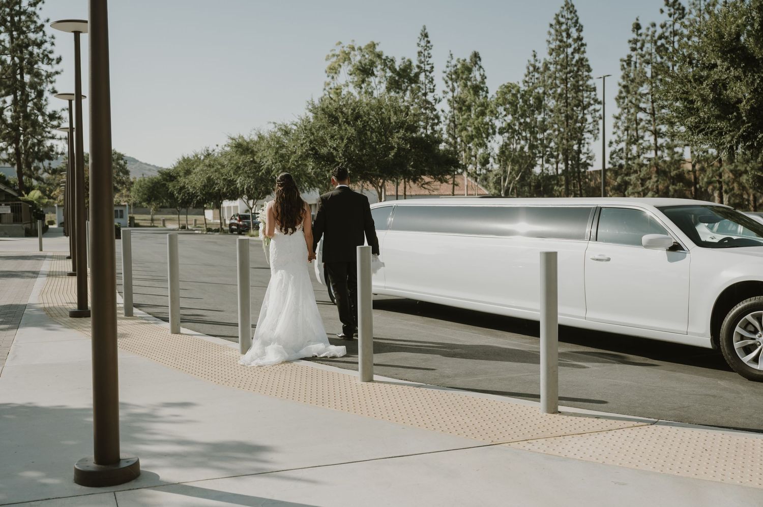 Bride and groom walking hand in hand toward a white stretch limousine on their wedding day.