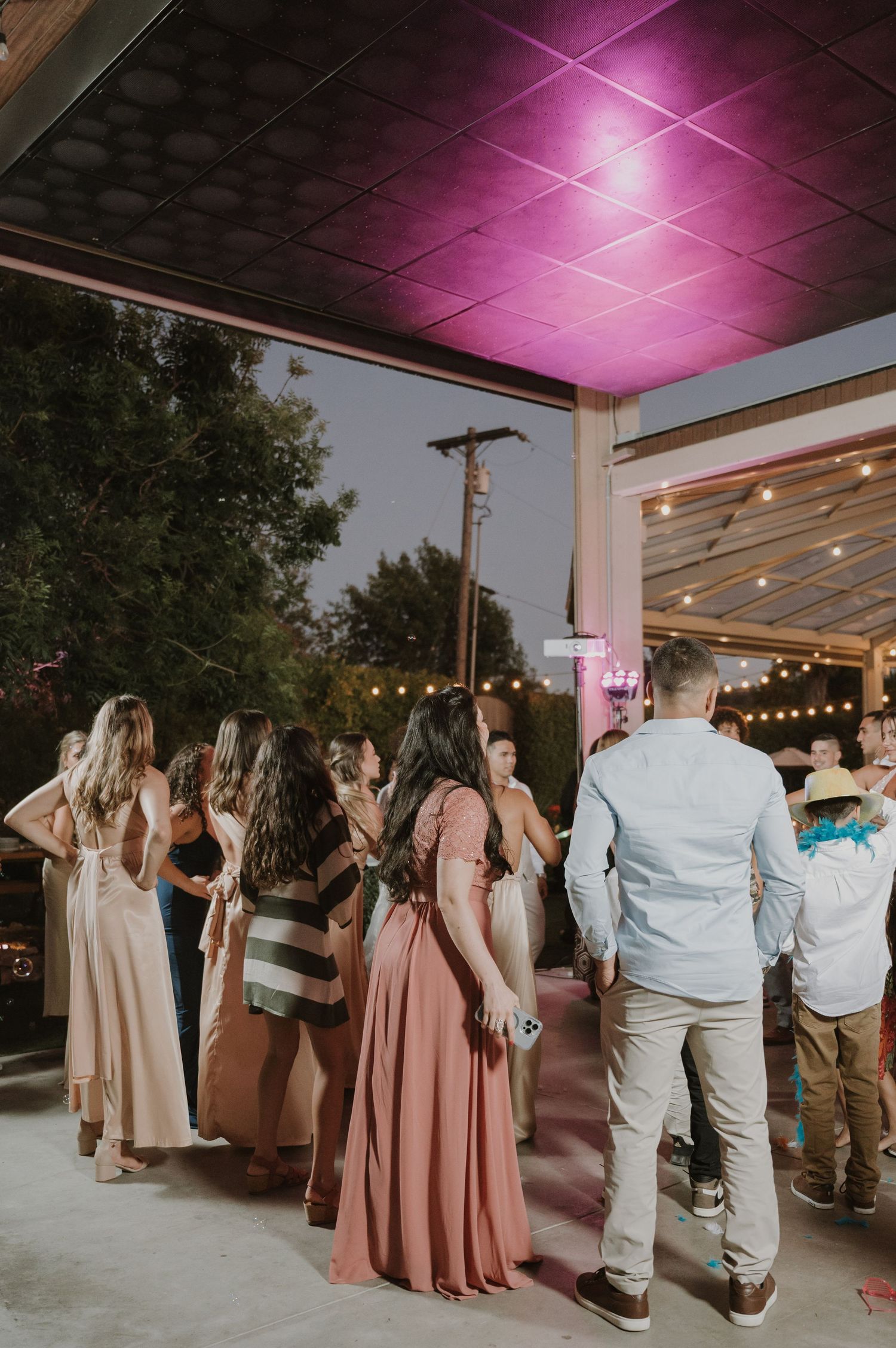 Guests dancing outdoors at a wedding reception under pink-lit ceiling with string lights at night.