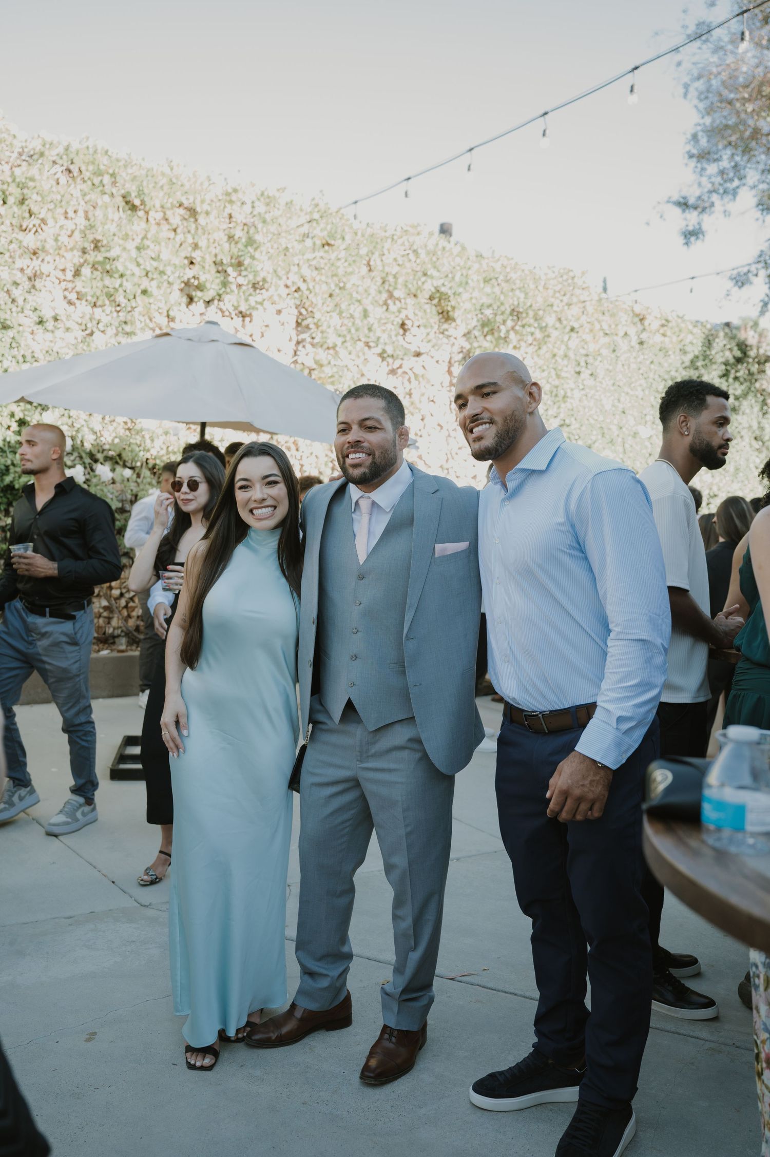 A woman in a blue gown poses with two men in suits at an outdoor wedding reception.