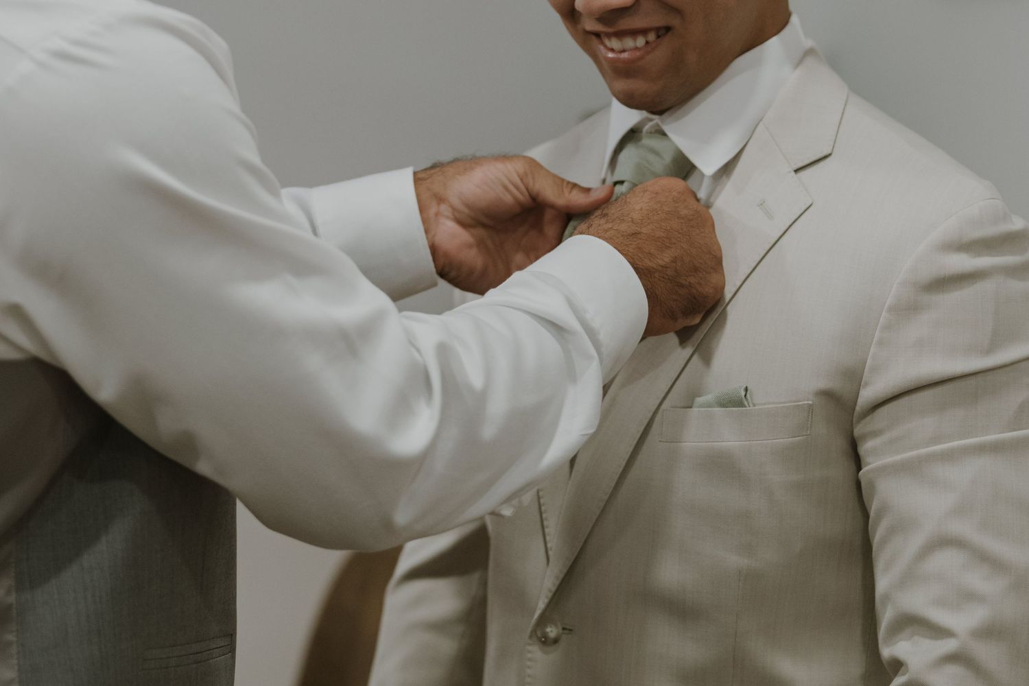 Close-up of groomsman adjusting sage green tie on smiling groom in elegant ivory wedding suit.
