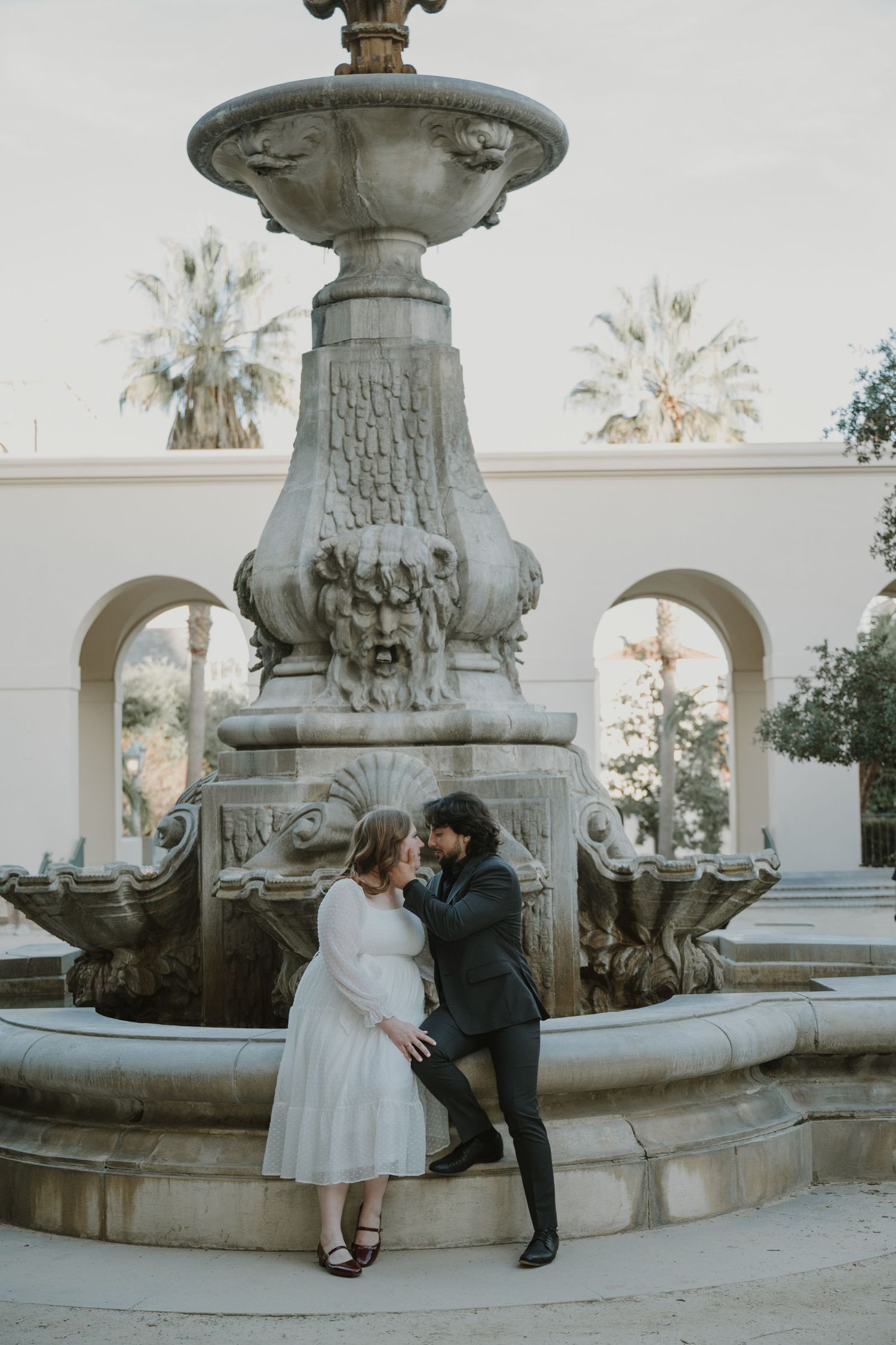 Couple sharing an intimate moment by an ornate stone fountain in a classical courtyard setting.