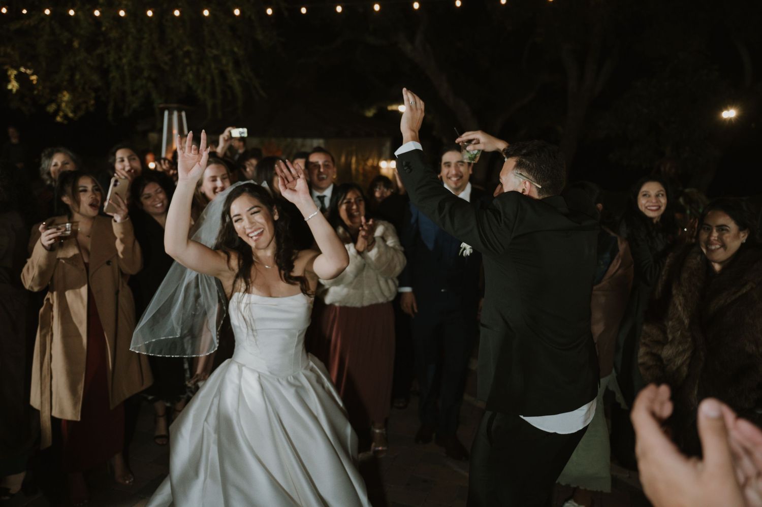 Wedding party dancing under string lights at an outdoor evening celebration.
