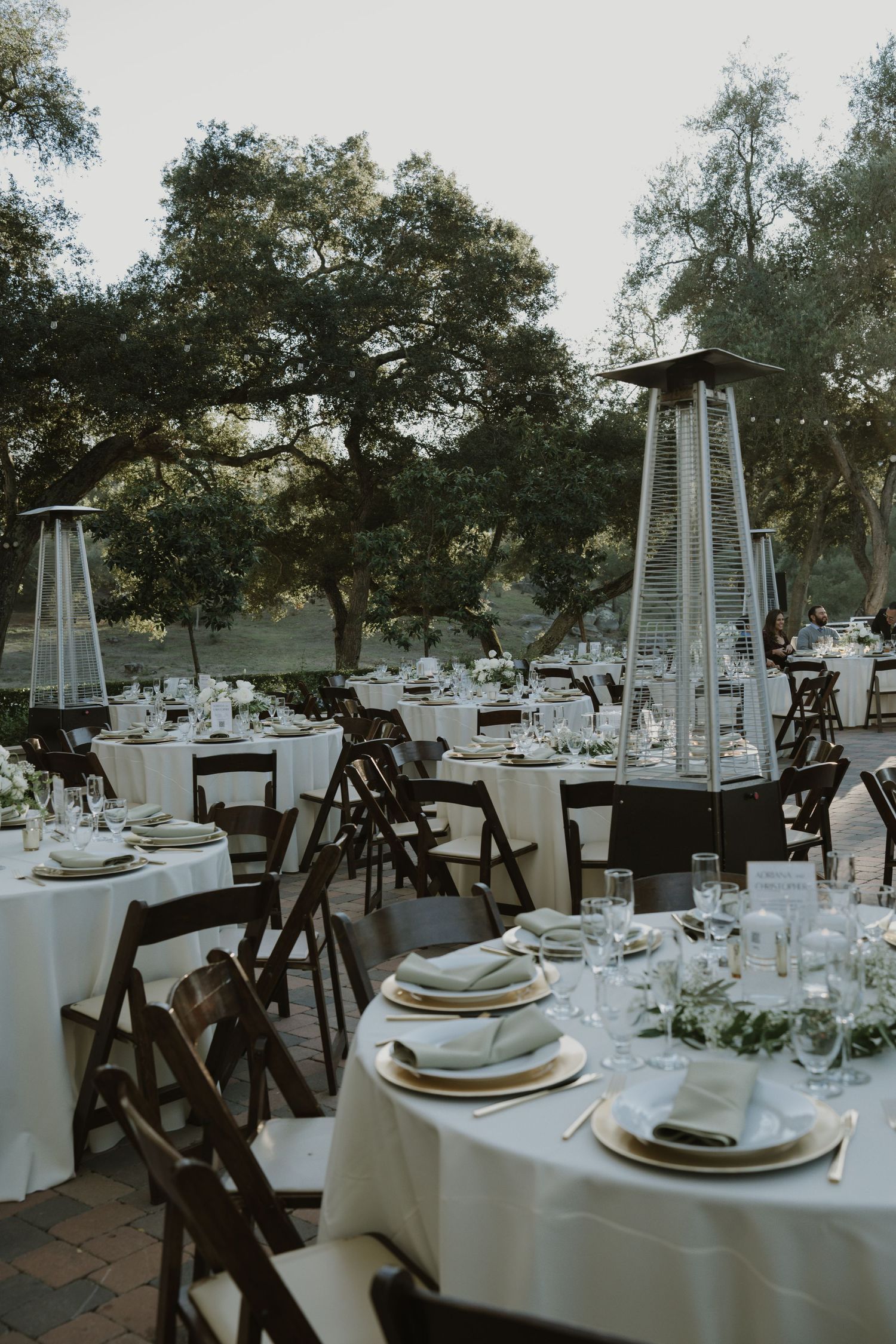 Outdoor wedding reception tables set under oak trees with white tablecloths and dark wooden chairs.