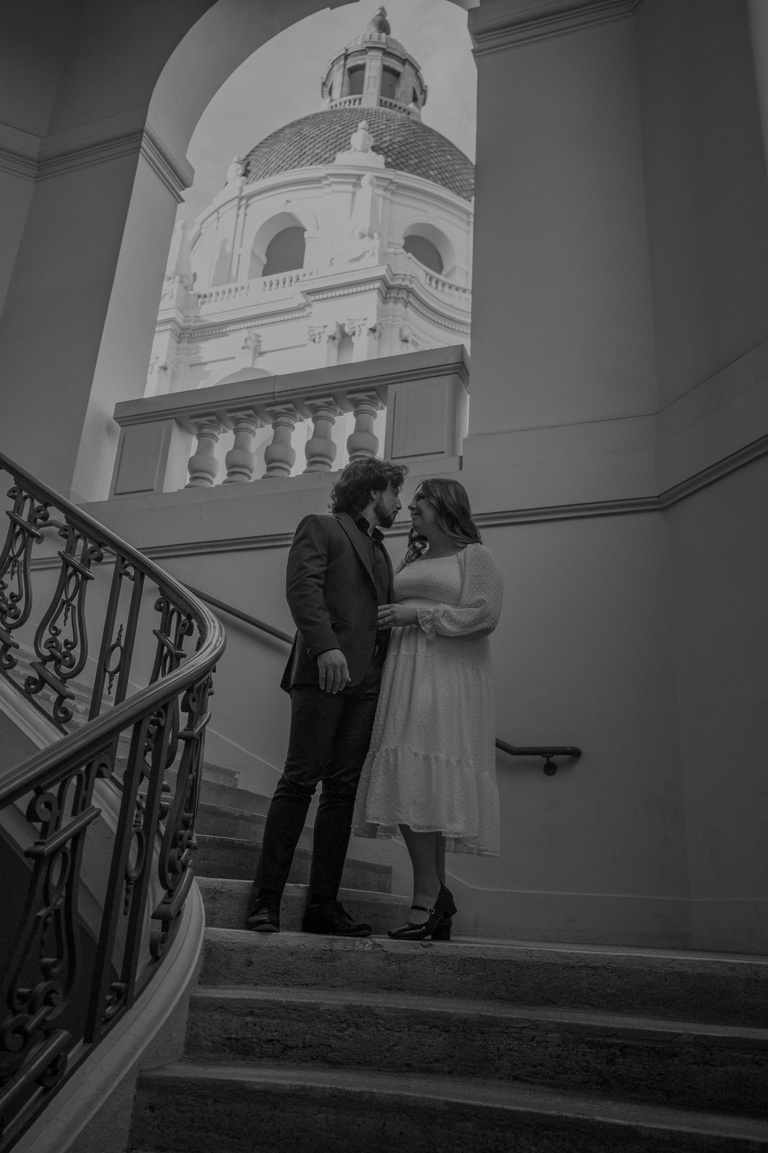 Couple poses on elegant staircase with ornate iron railing and domed architecture.
