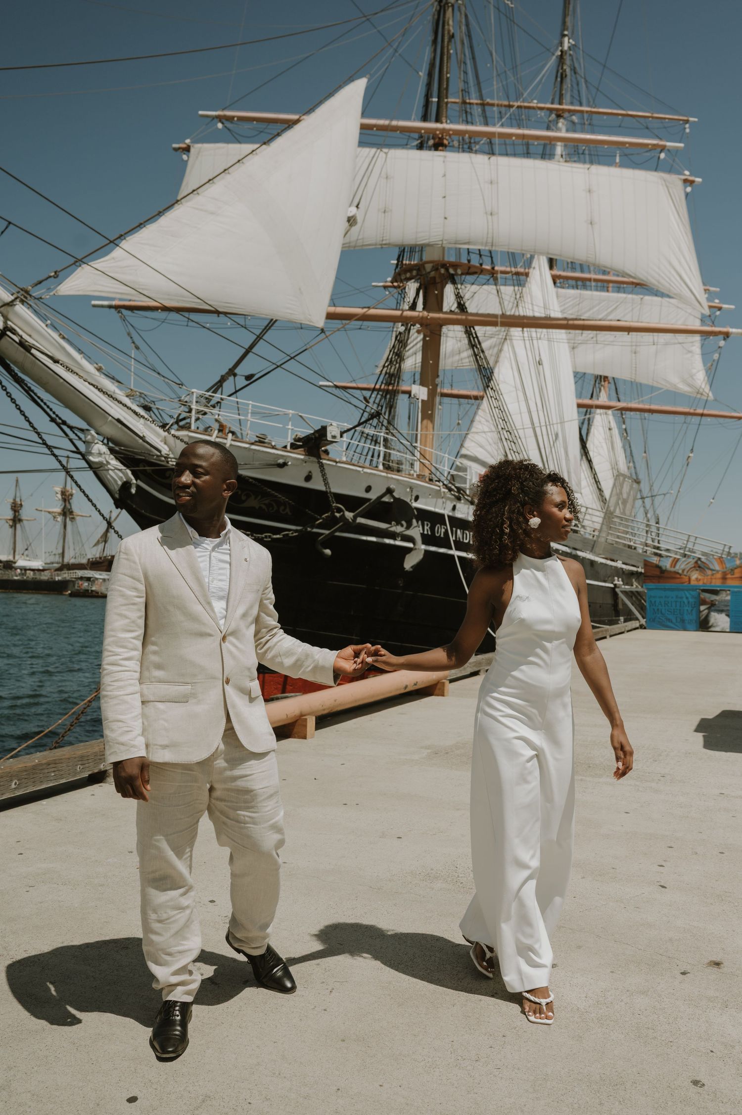 A couple in all-white outfits poses in front of a tall ship at a harbor.