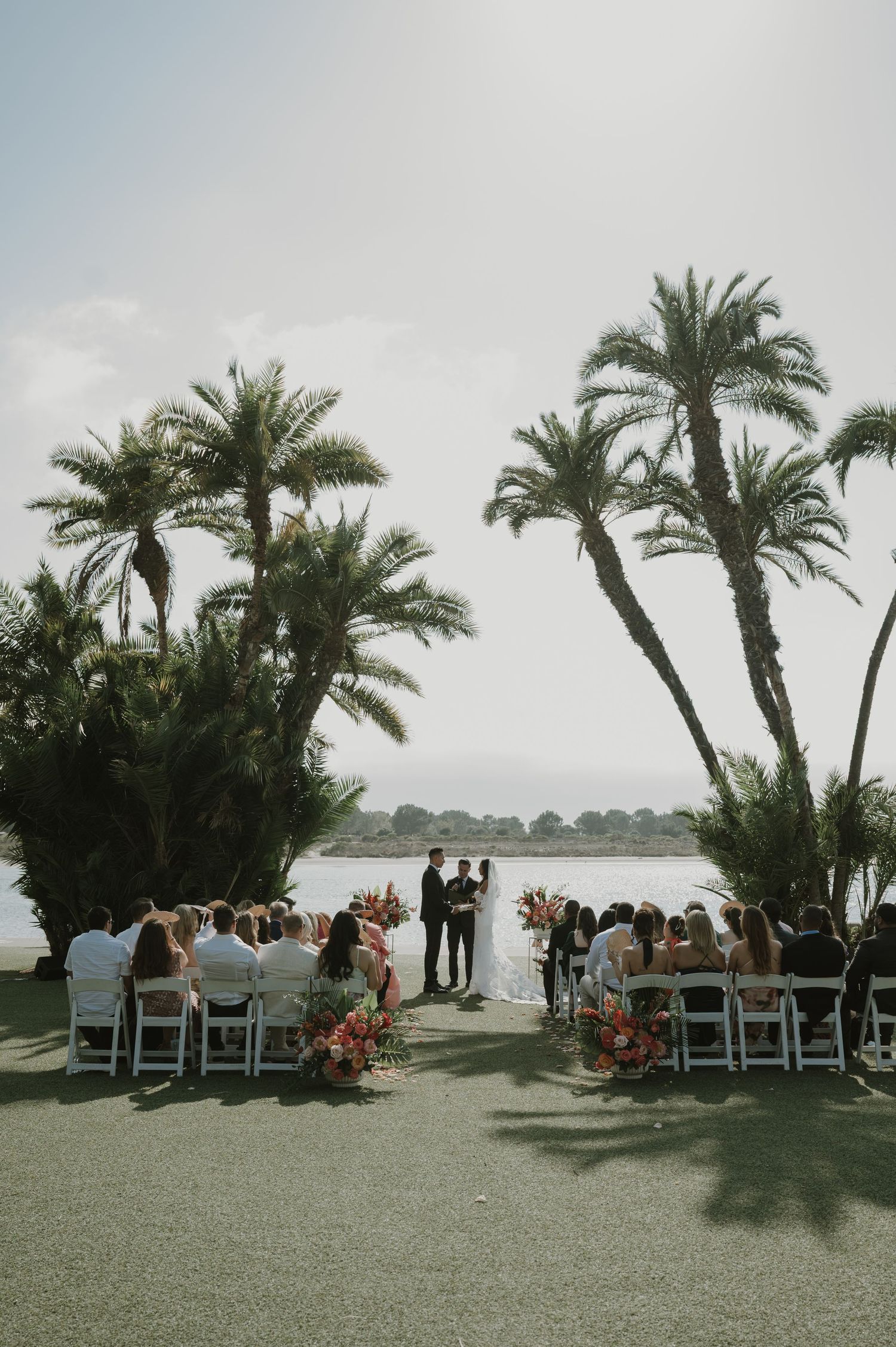 Wedding guests seated in white chairs under palm trees at an oceanside ceremony.