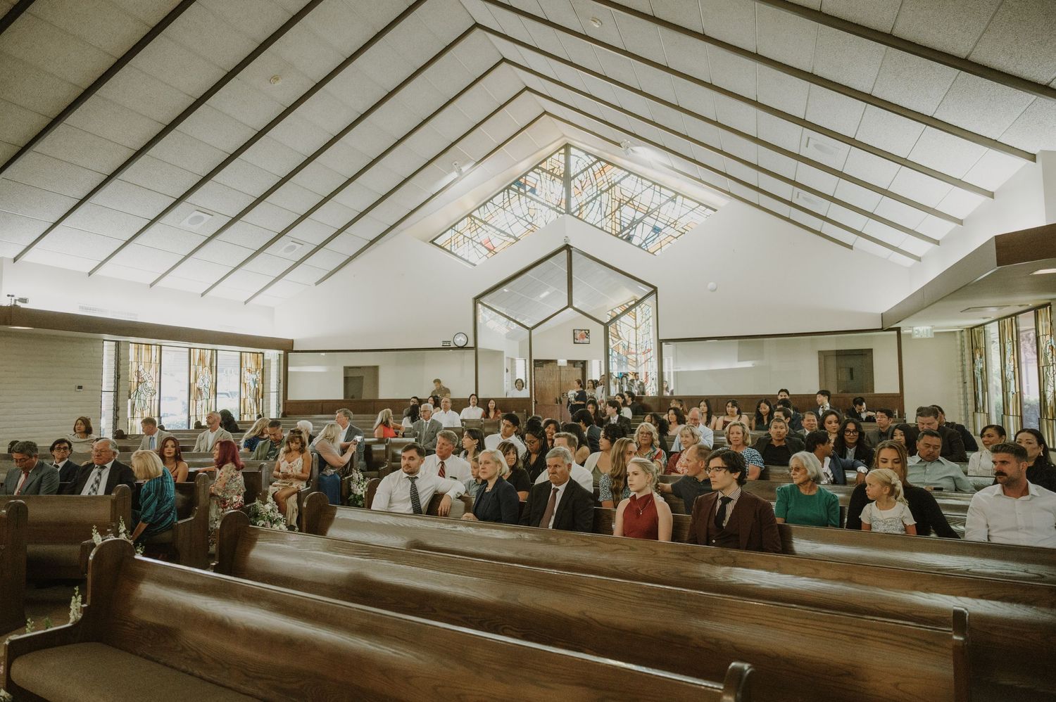 Guests seated in wooden church pews during a wedding ceremony in a bright chapel.