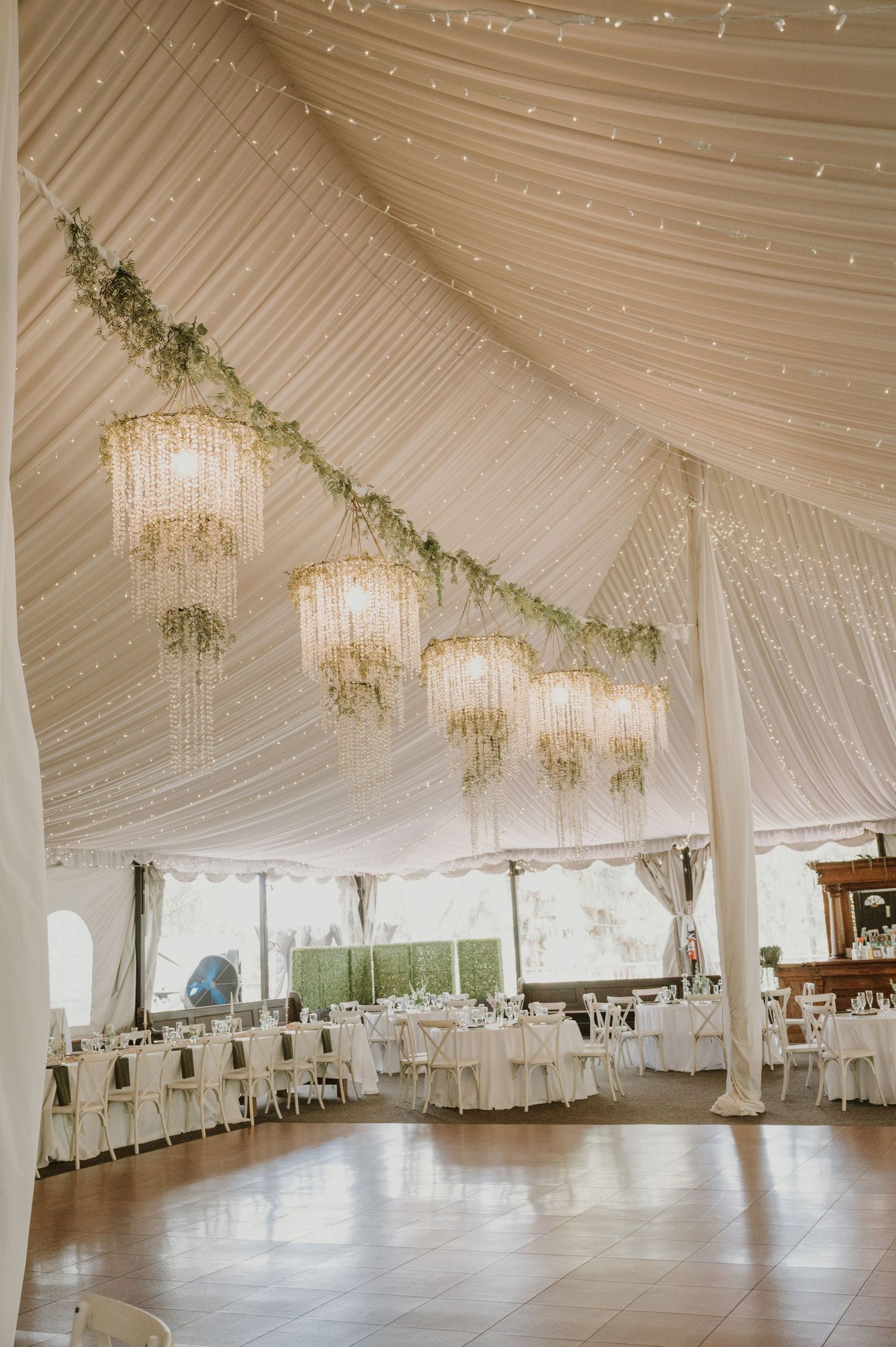 Elegant wedding reception tent with crystal chandeliers, fairy lights, and greenery garlands over white-draped tables.