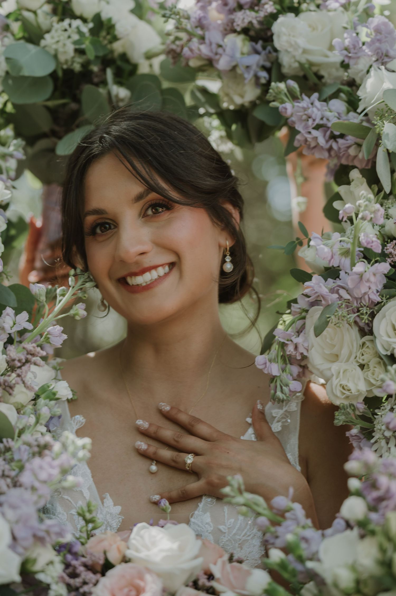 Smiling bride surrounded by lush purple and white floral arrangement, wearing pearl earrings and ring, hand on chest.