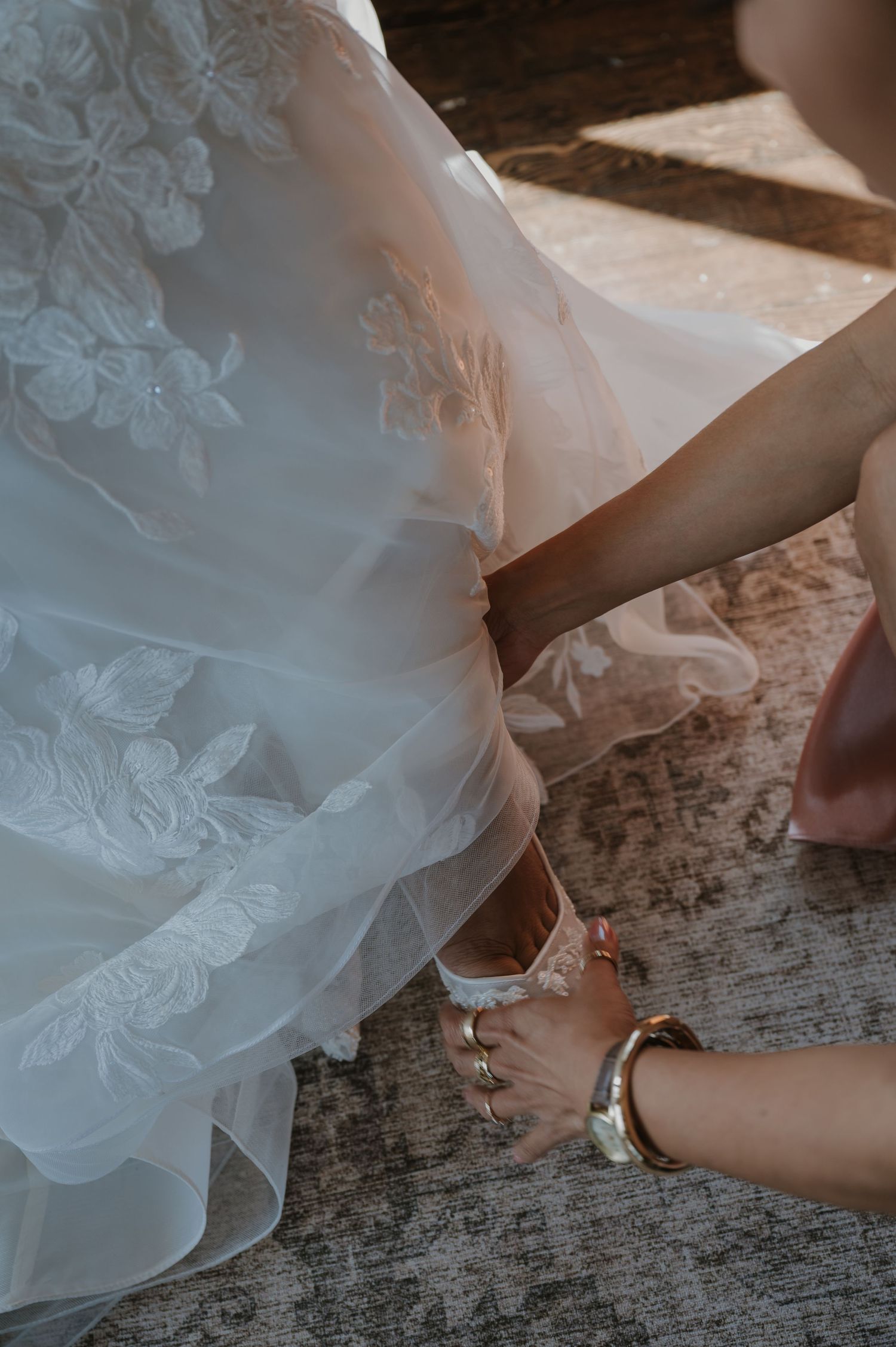 A helper fastens strappy silver heels on a bride wearing a white lace wedding gown.