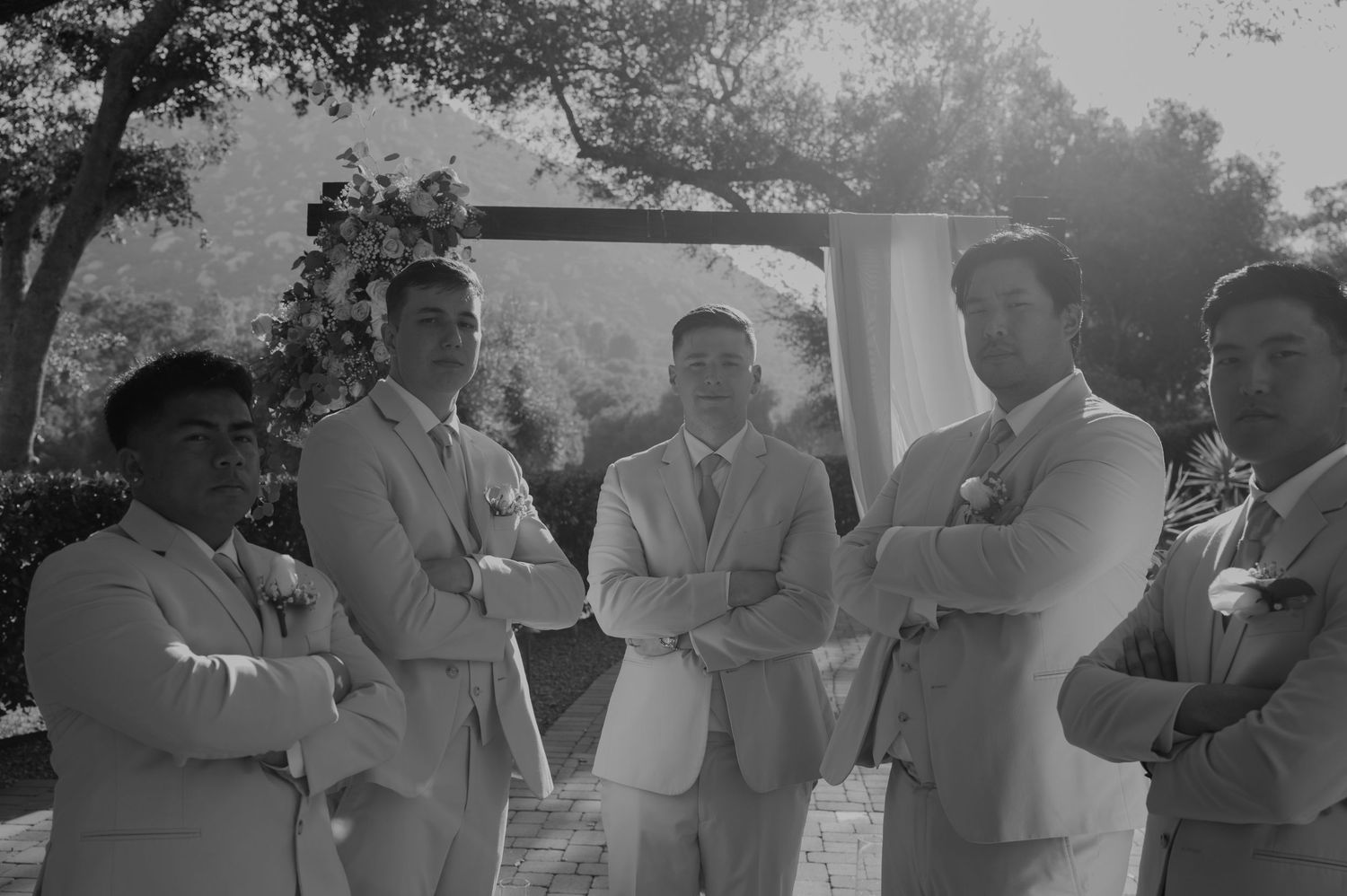 Five groomsmen in suits pose with arms crossed at outdoor wedding ceremony.