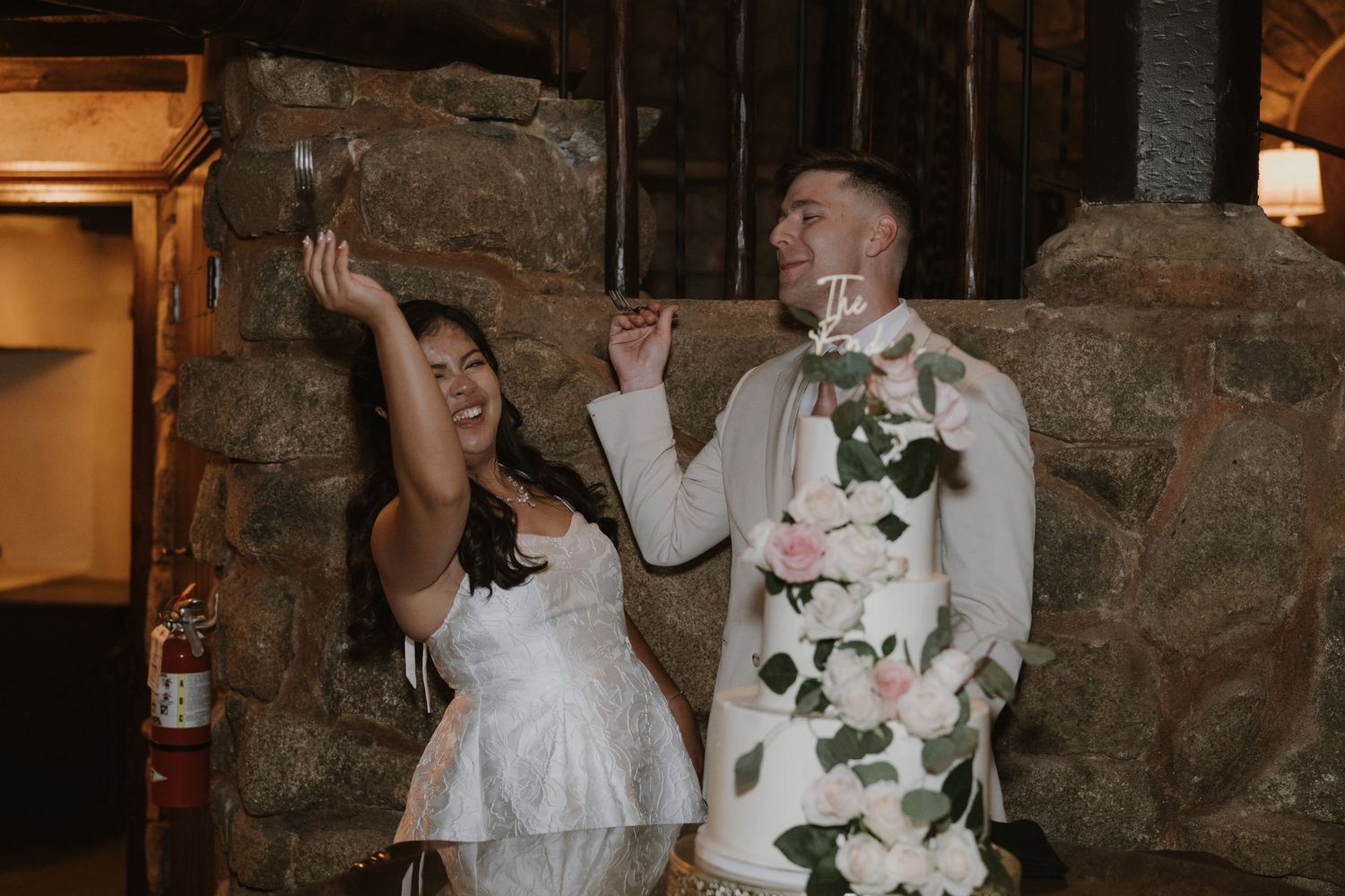 Bride in white dress and groom in white suit celebrating with drinks beside a floral wedding cake in a stone venue.