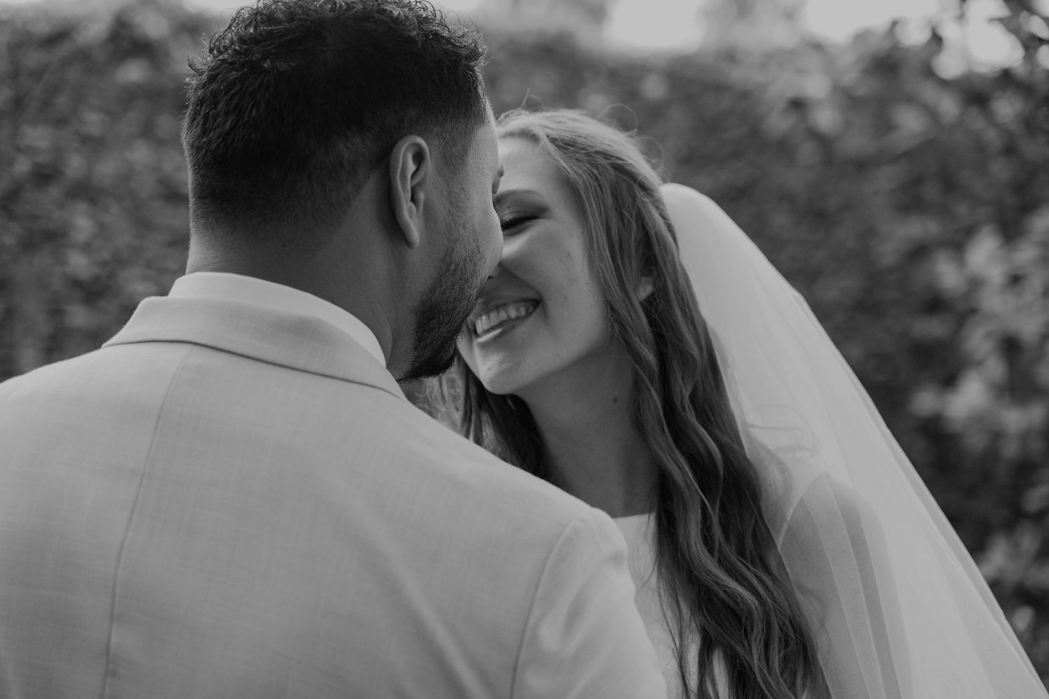 Black and white photo of smiling bride in veil sharing intimate moment with groom at outdoor wedding ceremony.