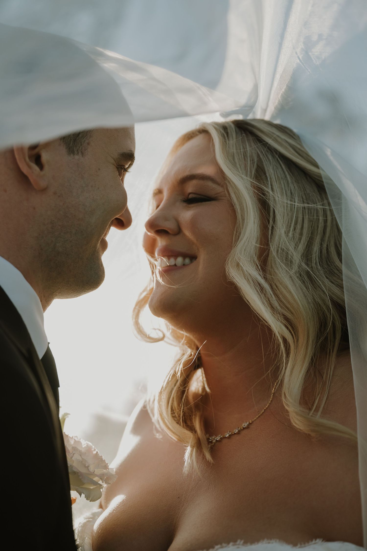 Bride and groom share an intimate moment under a flowing veil, bathed in golden backlight on their wedding day.