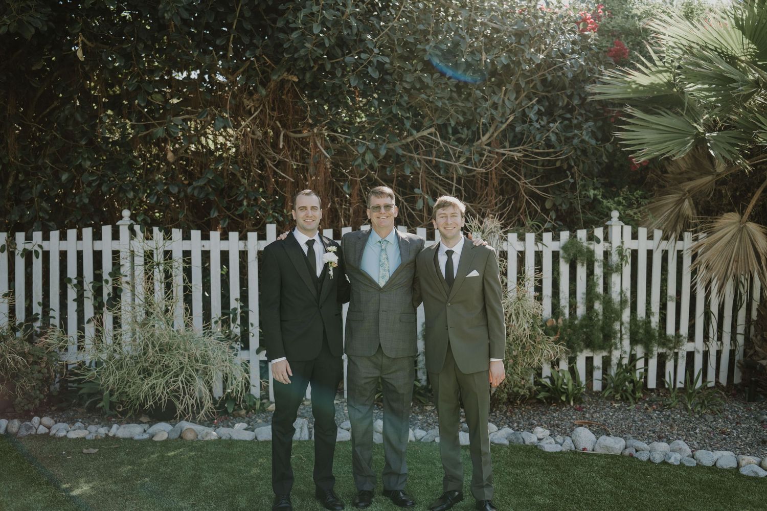 Groom and groomsmen in dark suits pose by white picket fence at outdoor wedding.