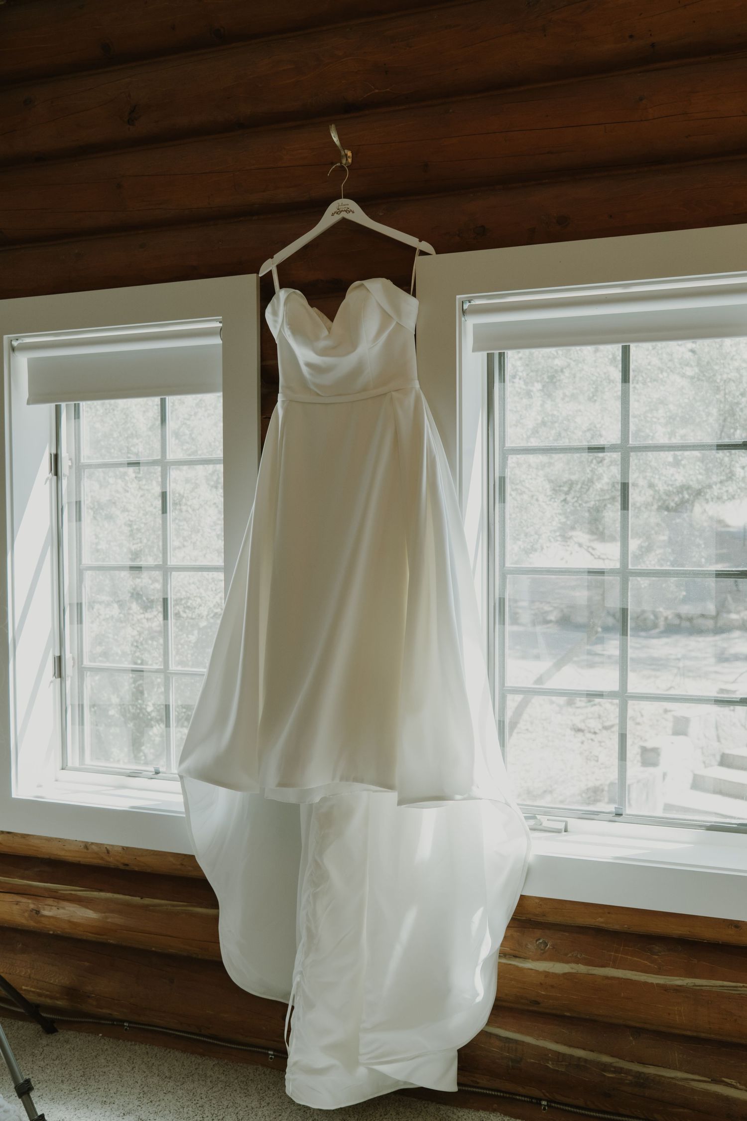 Elegant white strapless wedding dress with a sweetheart neckline hanging in a rustic log cabin room with large windows.