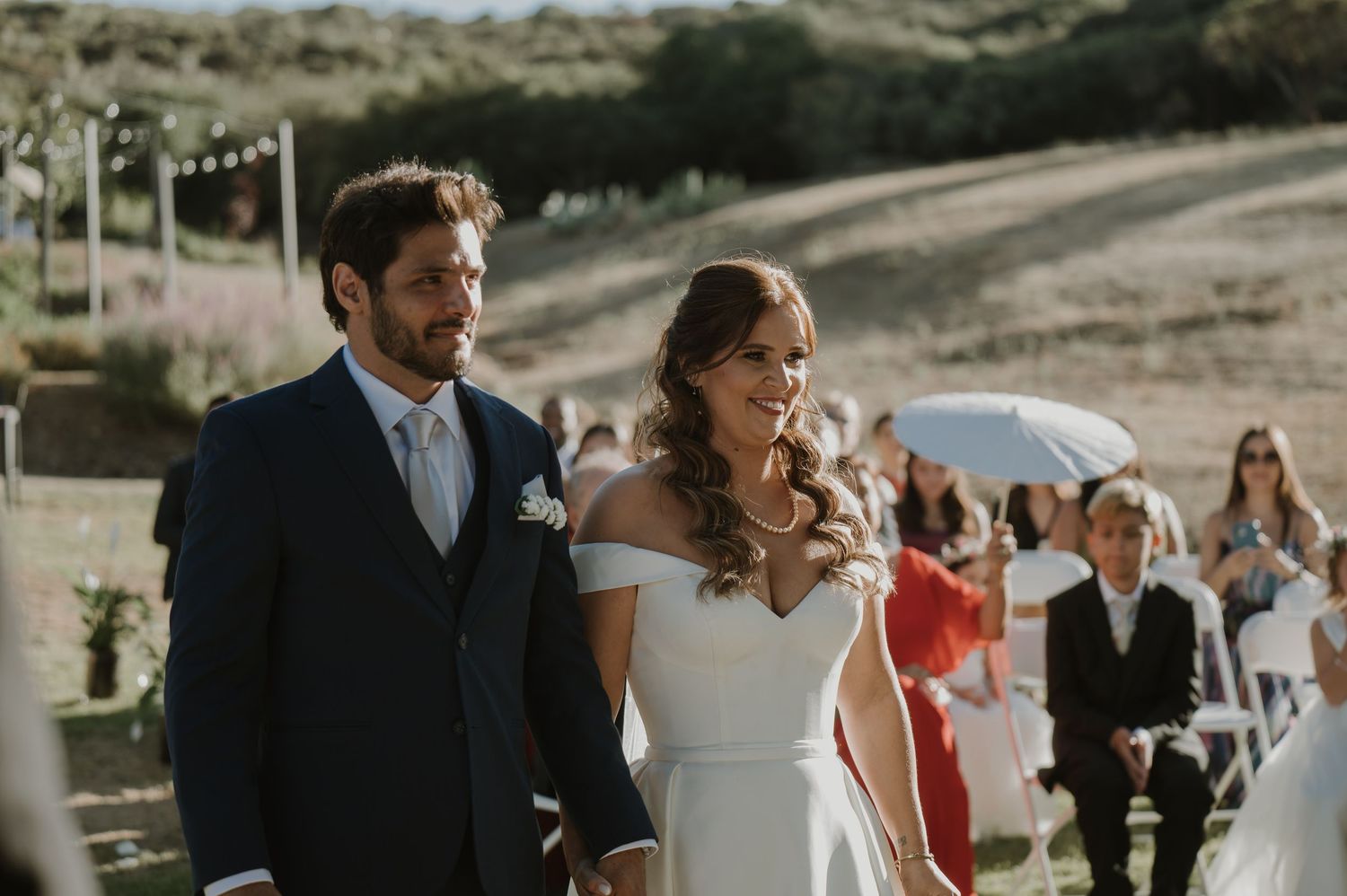Newlywed couple walks hand in hand down aisle after outdoor wedding ceremony.