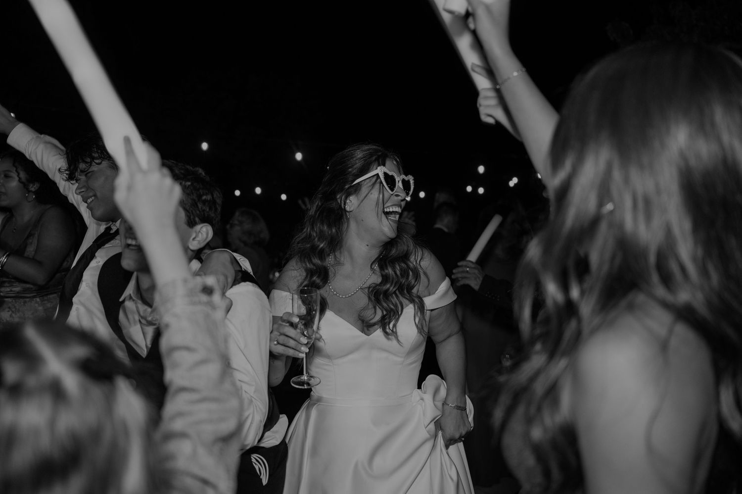 Bride in white dress and oversized sunglasses dancing joyfully at a wedding reception in black and white photo.