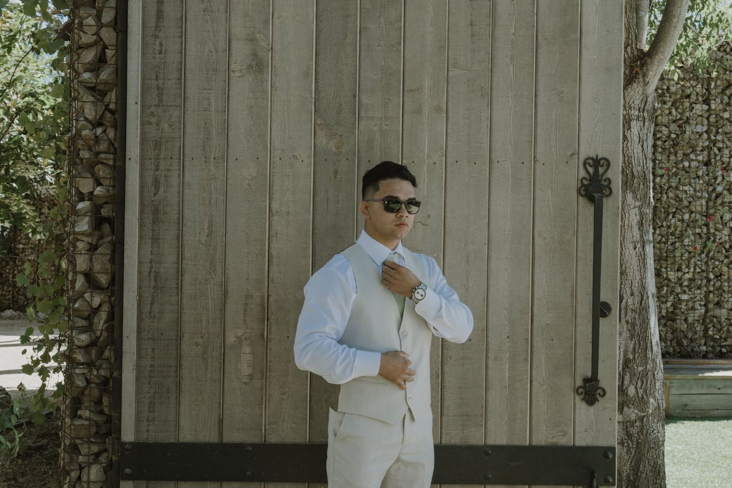 Stylish groom in a white suit and sunglasses adjusting his jacket outside rustic barn doors.