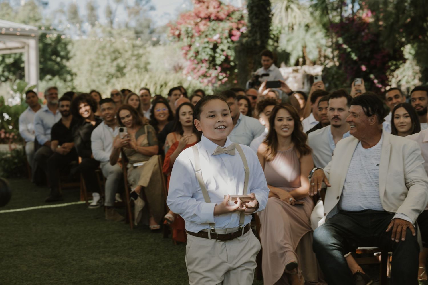 A young boy walks down the aisle at an outdoor wedding ceremony while guests smile and watch from their seats.