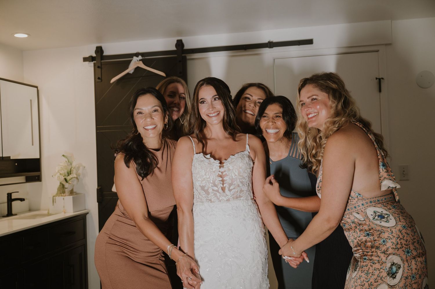 Bride in lace wedding gown smiling with bridesmaids in front of barn door during wedding preparations.
