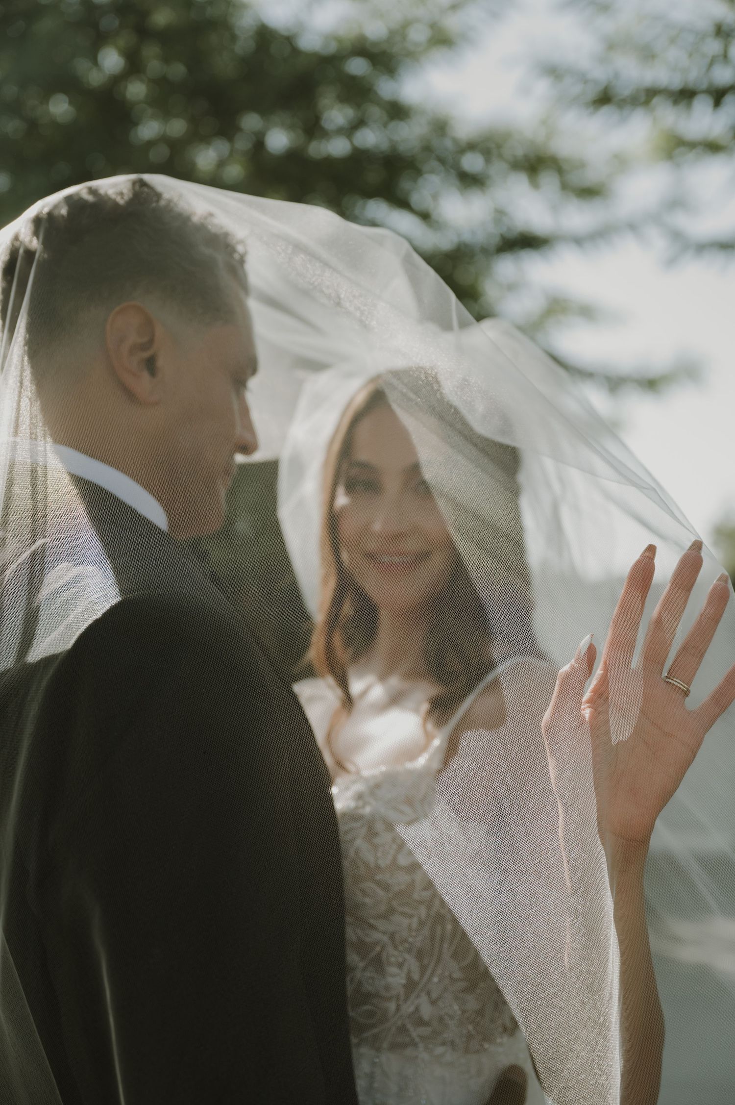 Bride smiling under a sheer veil beside groom, showing off her wedding ring in soft natural light.