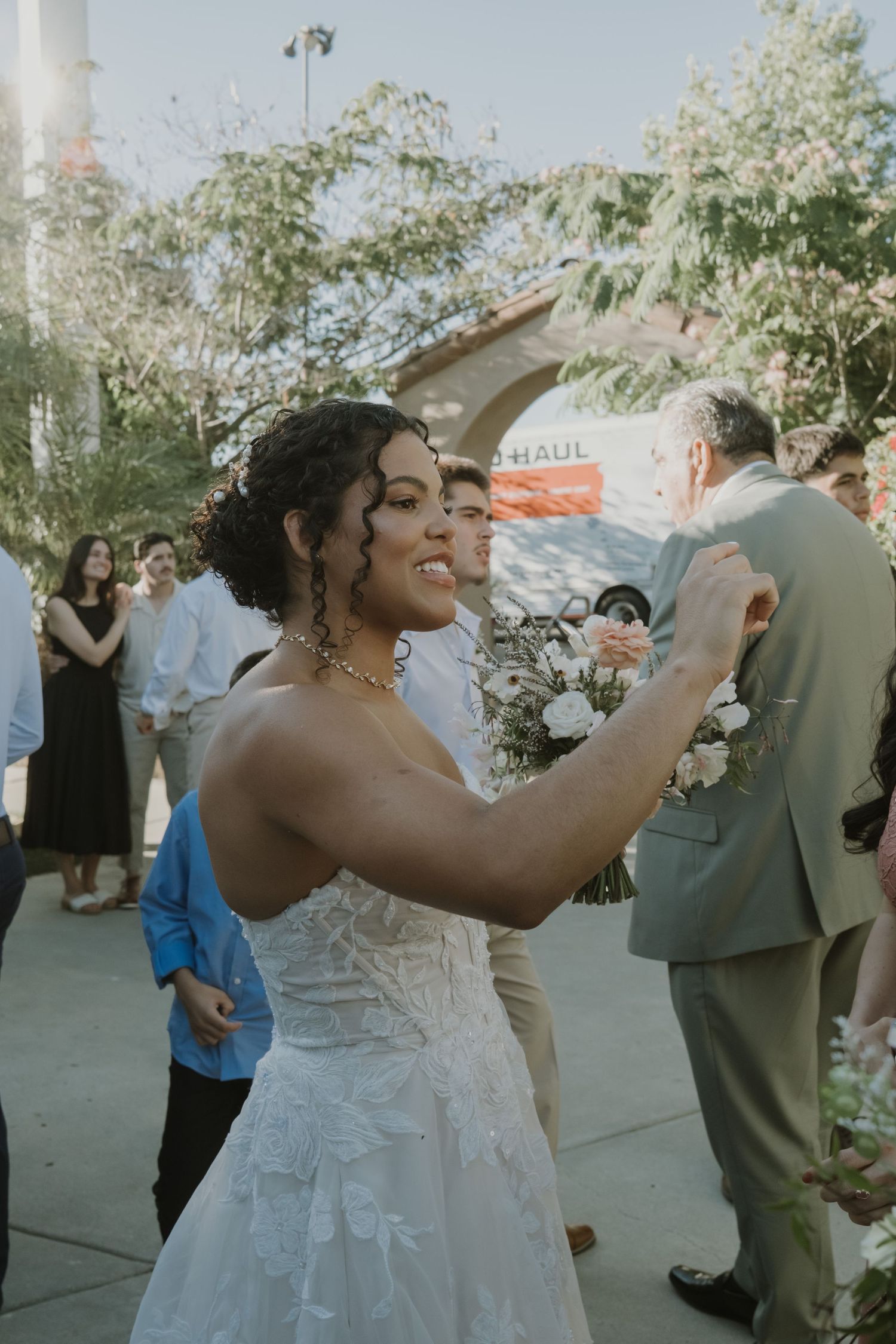 A bride in a white lace gown dances joyfully with a guest during an outdoor wedding celebration.