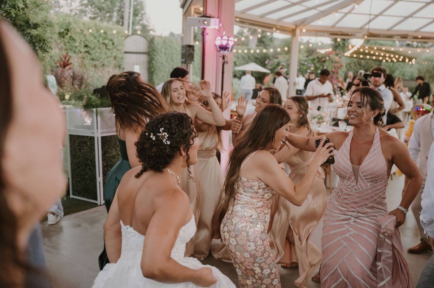Bride and guests dancing joyfully at an outdoor wedding reception under a tent with string lights.