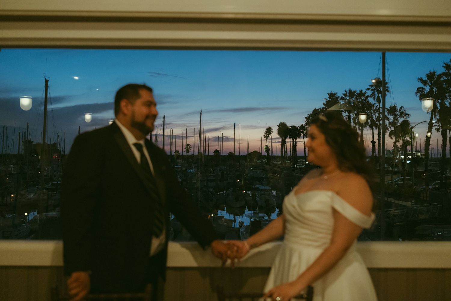 Bride and groom hold hands at sunset overlooking a marina with palm trees at dusk.