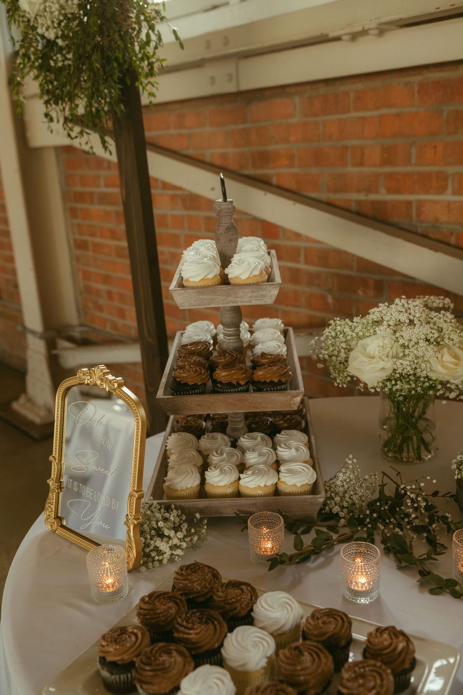 Elegant tiered cupcake tower display with white and chocolate frosted cupcakes, candles, and floral arrangements at a wedding reception.