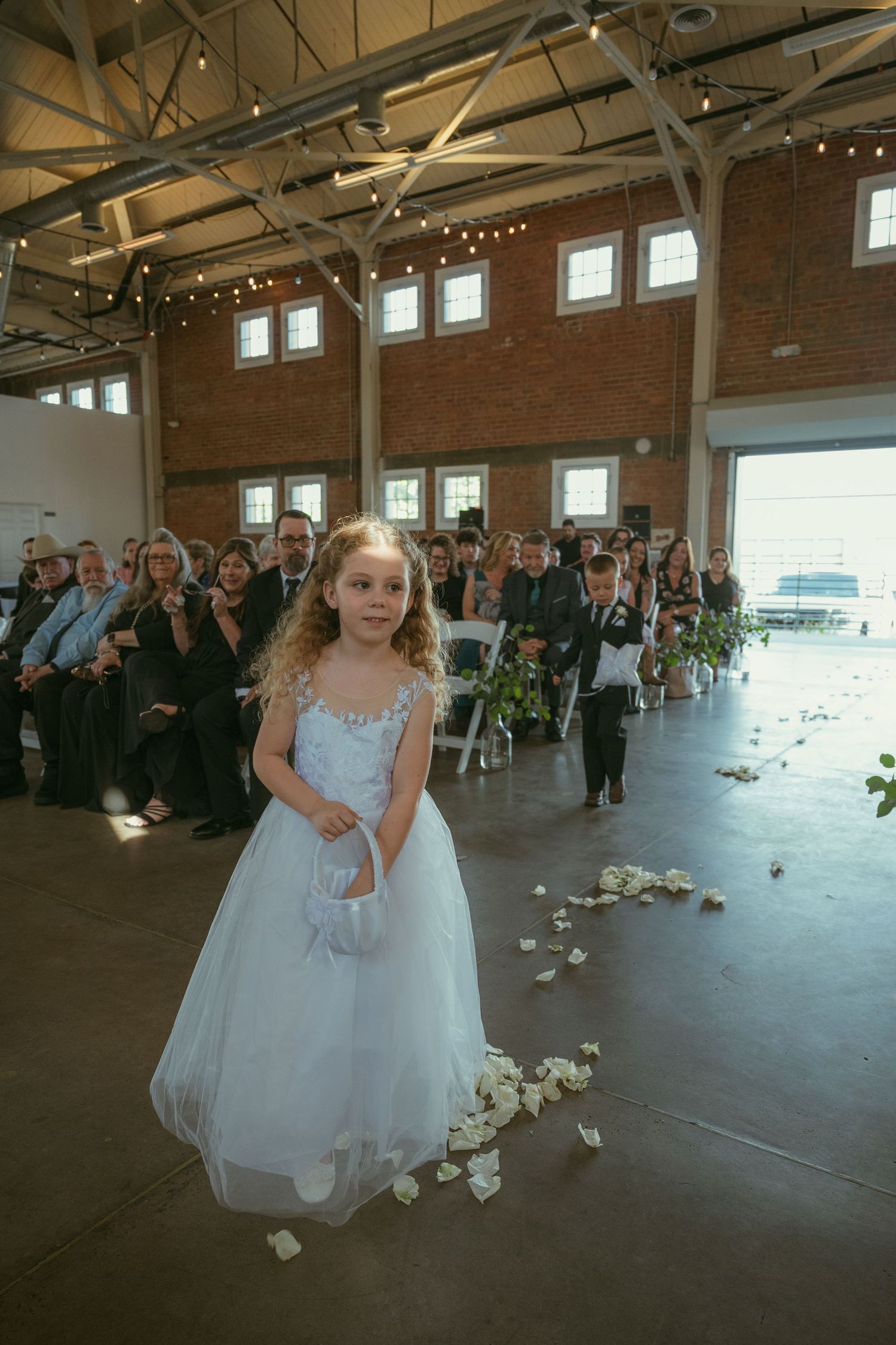 A flower girl in a white dress scatters rose petals down the aisle at an indoor wedding ceremony.