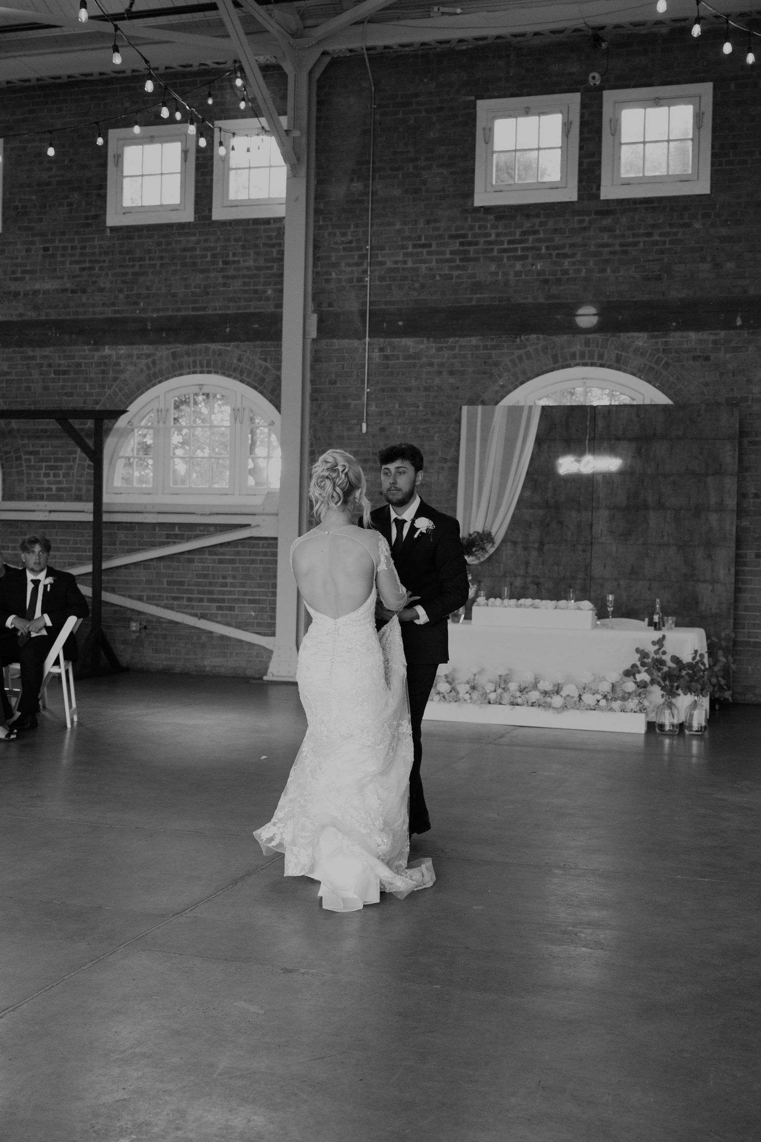 Bride and groom share their first dance in a rustic brick venue with string lights in black and white.