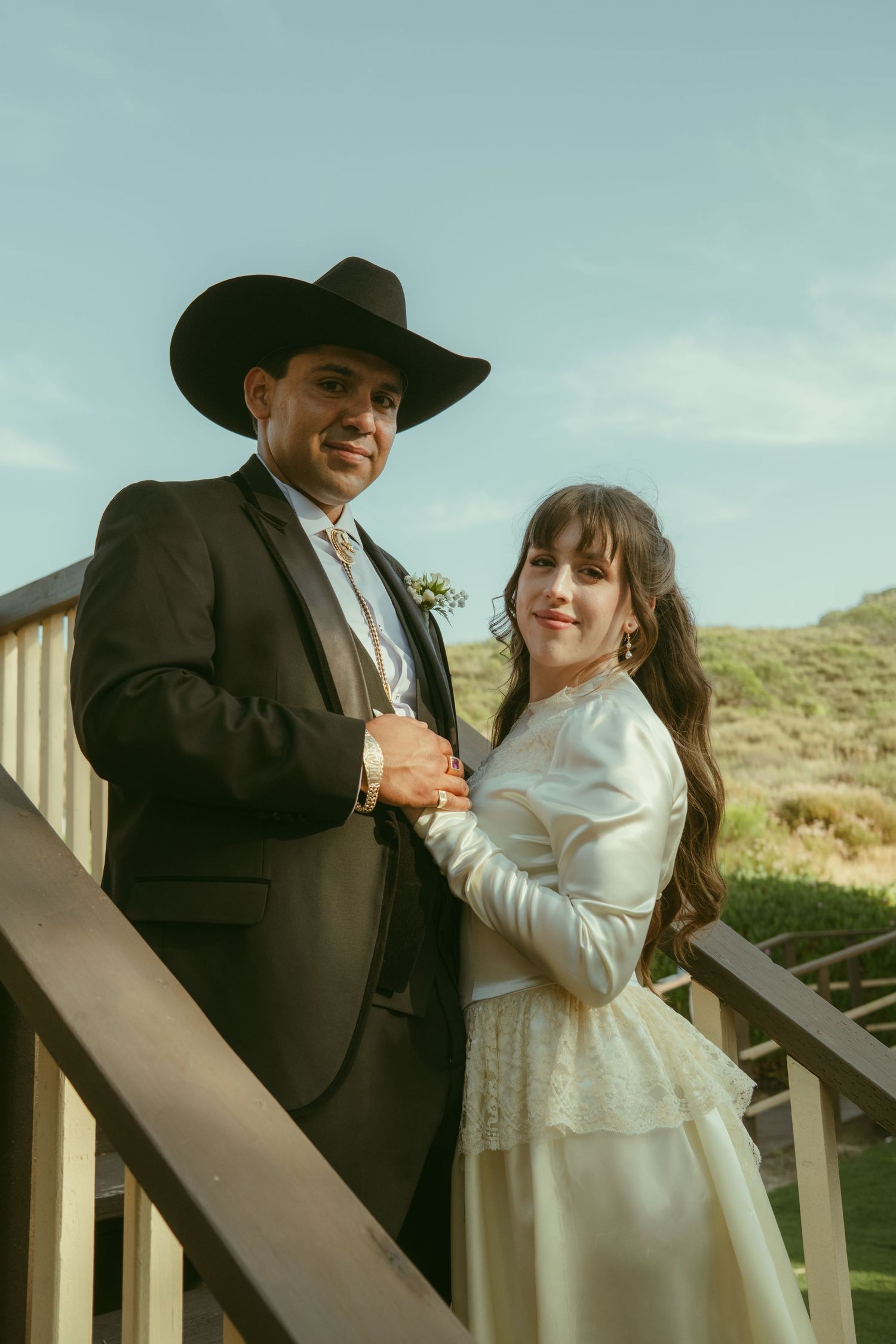 A couple in western wedding attire pose on outdoor stairs, groom in black cowboy hat and suit, bride in white lace dress.