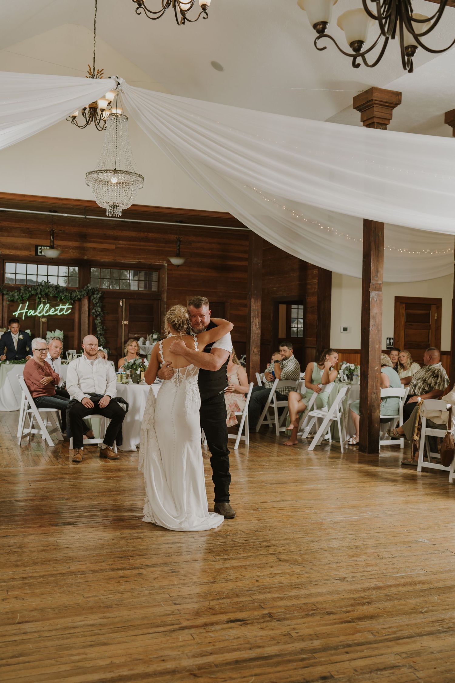 Wedding reception dance floor in rustic venue with white draping and chandeliers.