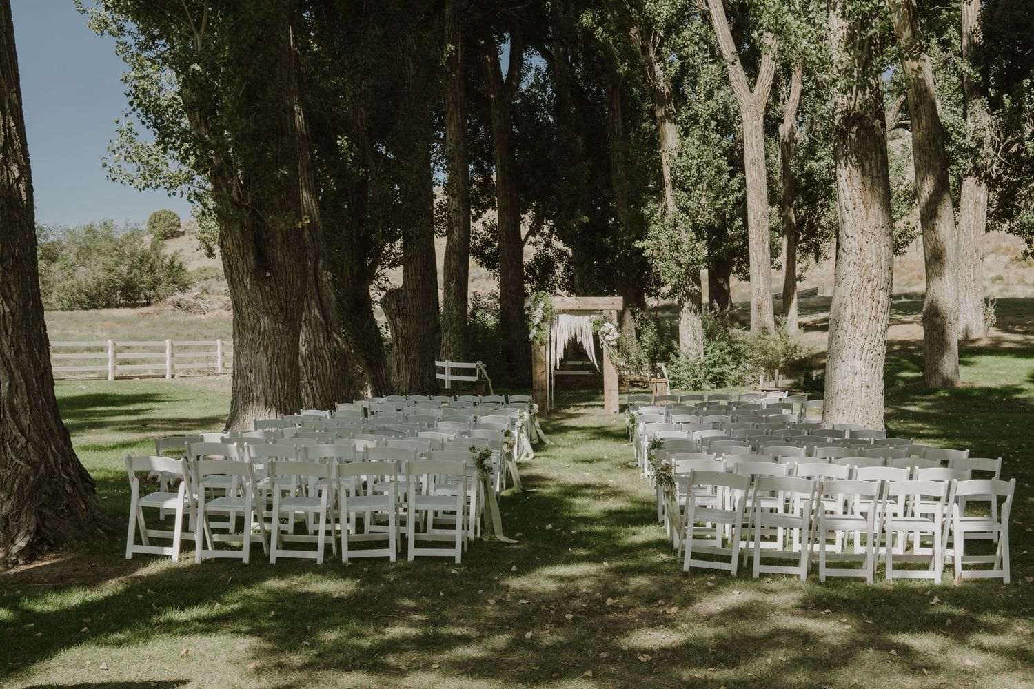 Outdoor wedding ceremony setup with white chairs arranged under large shady trees.