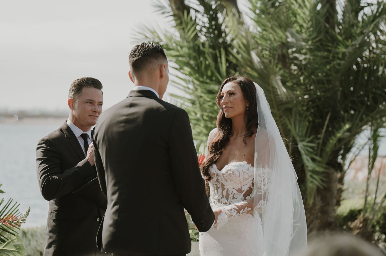 Intimate wedding ceremony moment captured under swaying palm trees by the water.