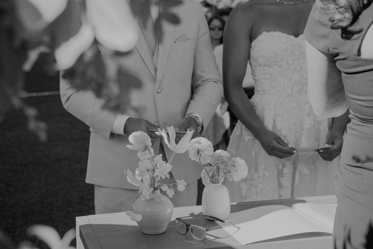 Black and white wedding ceremony photo of couple at altar with floral arrangements on table.