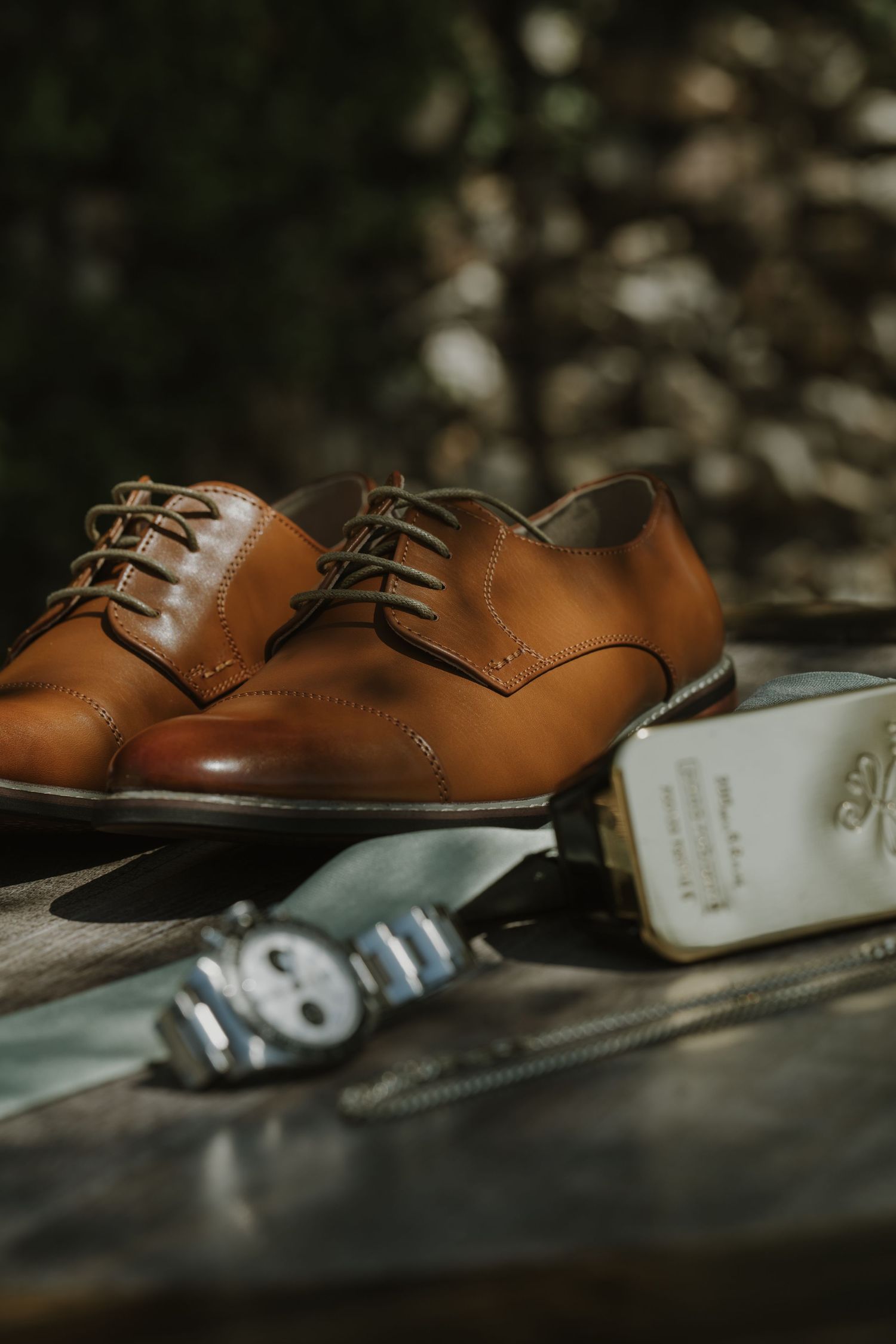 Polished tan leather Oxford dress shoes beside a watch and white book on a dark surface.