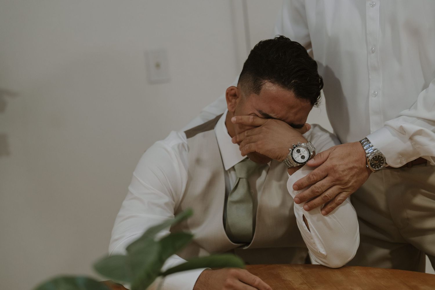 Emotional groom covering his face while someone helps him with his tie before the wedding.