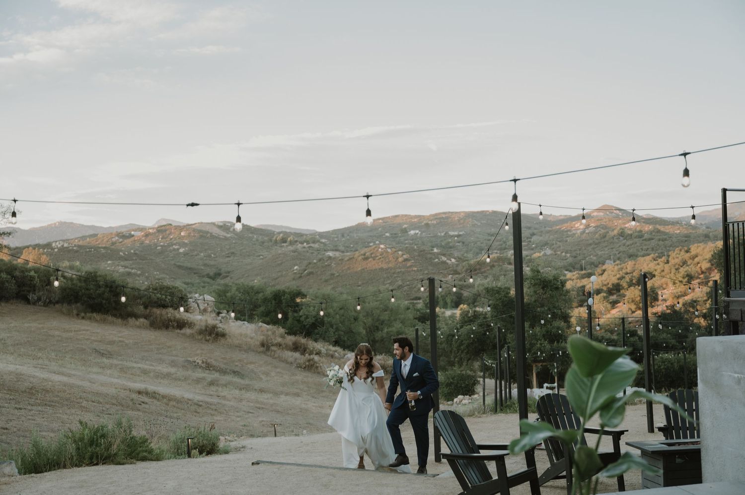 Bride and groom walk hand in hand at outdoor wedding venue with rolling hills and string lights backdrop.