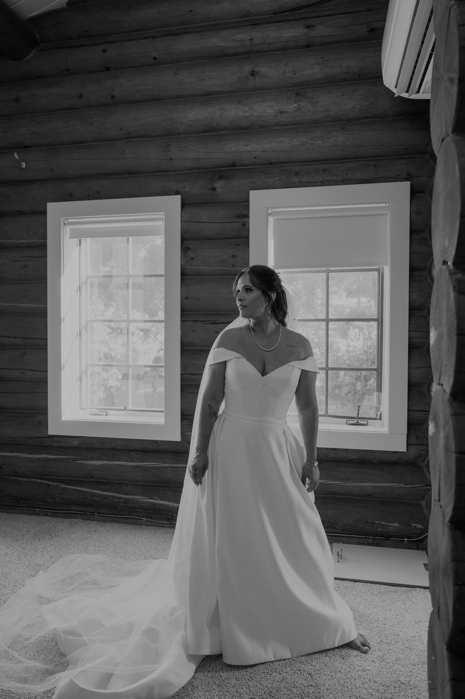 Elegant bride poses in off-shoulder wedding gown inside rustic log cabin venue.